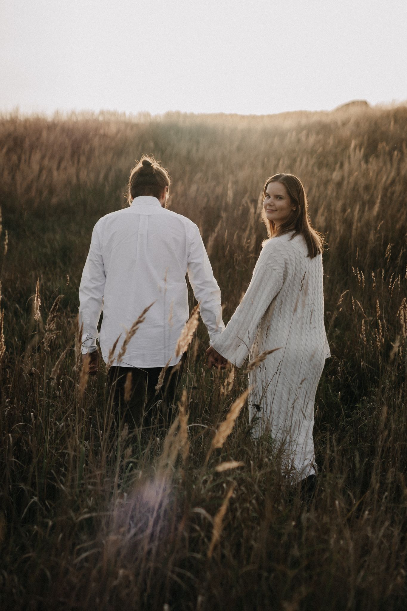 Couple walking hand in hand through tall golden grass field at sunset, woman glancing back smiling.