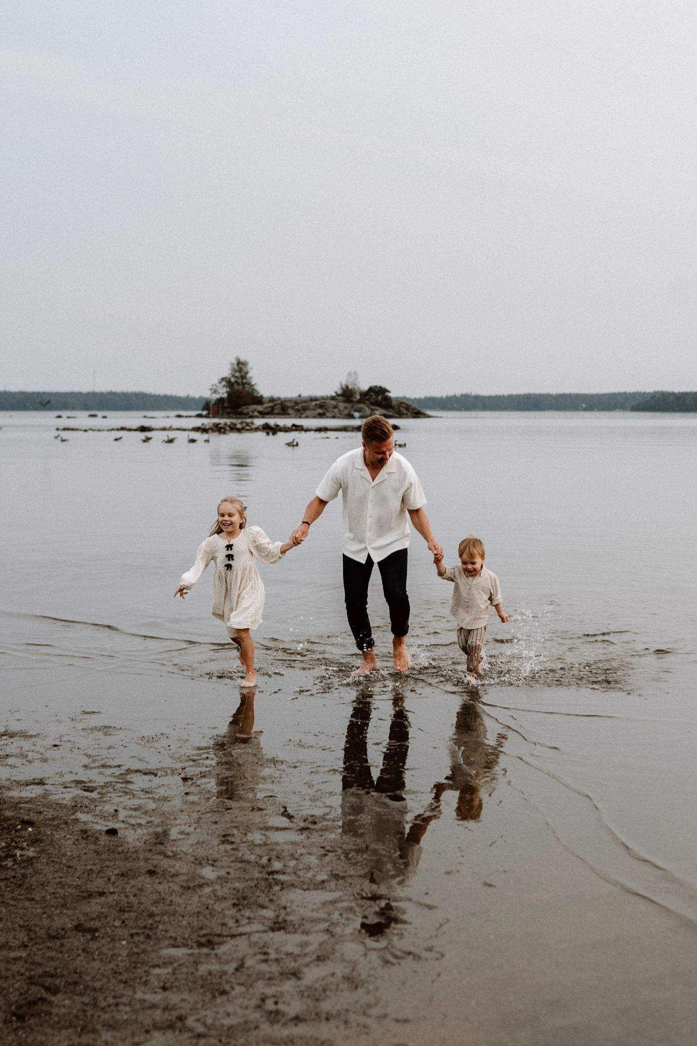Father holding hands with two young children, walking barefoot through shallow water on a beach.
