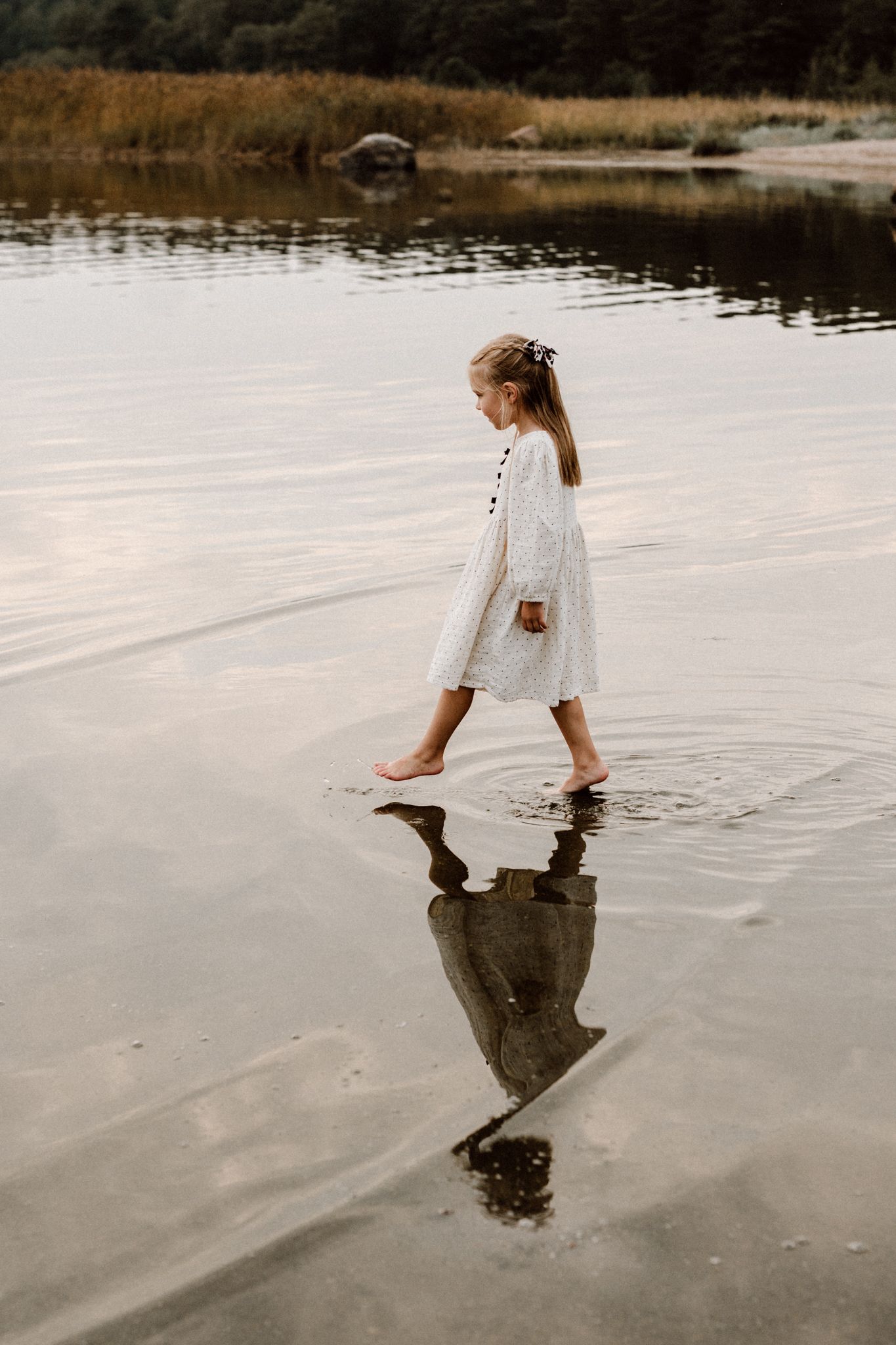 A young girl in a white dress walks barefoot through shallow water with her reflection visible below.