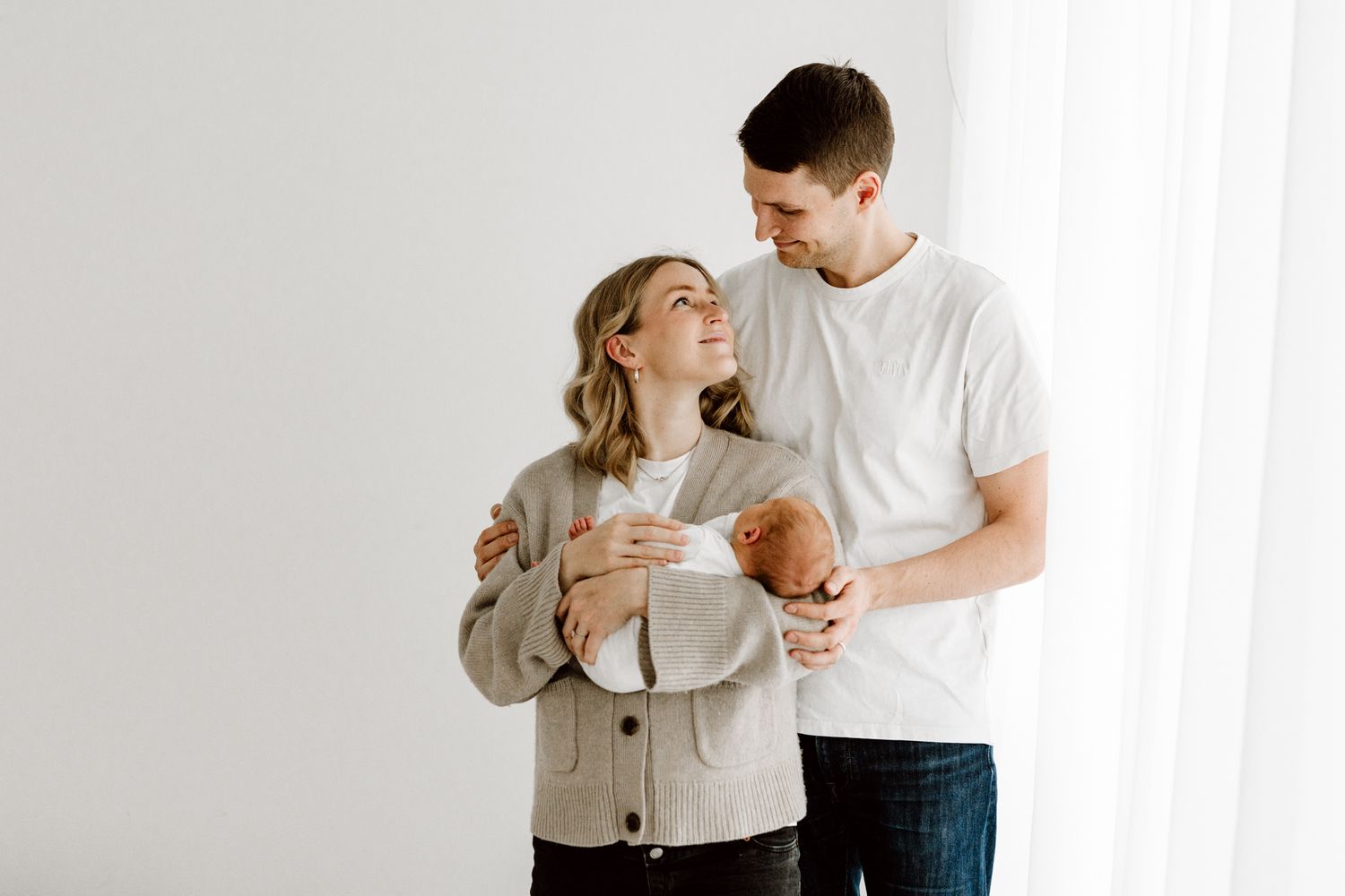 A smiling couple stands by a bright window, lovingly holding their newborn baby together.