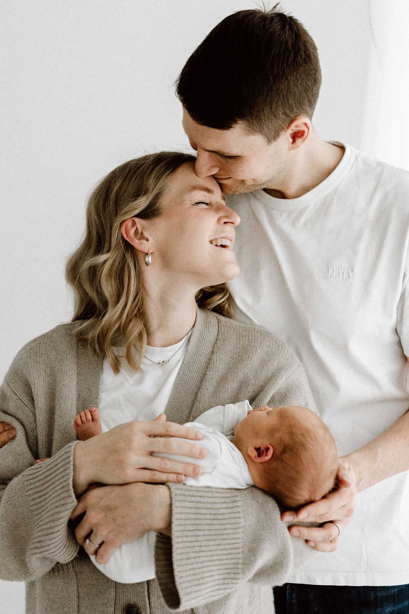 Happy parents smile and nuzzle together while holding their newborn baby in a bright white room.