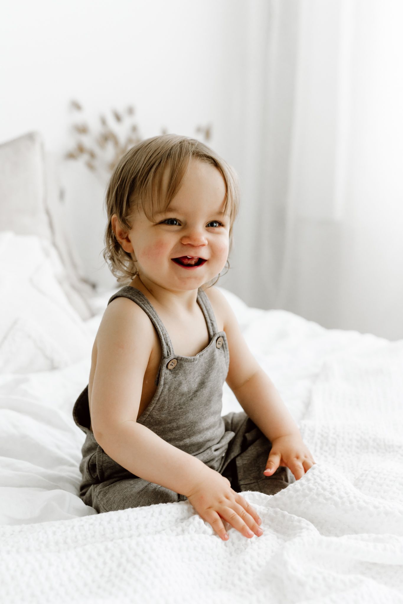 Adorable smiling toddler in gray overalls sitting on white bed in bright room.