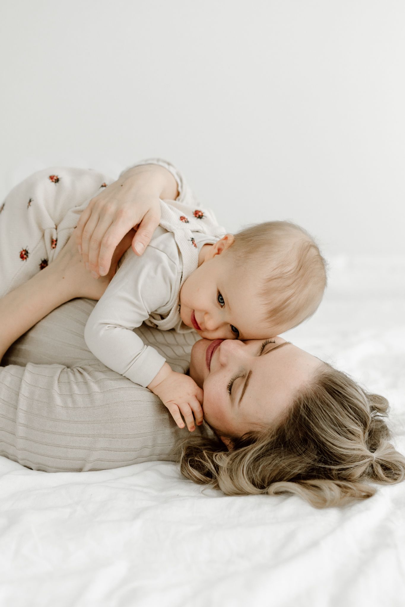 Mother lying on a white bed smiling as her baby leans in for a kiss.