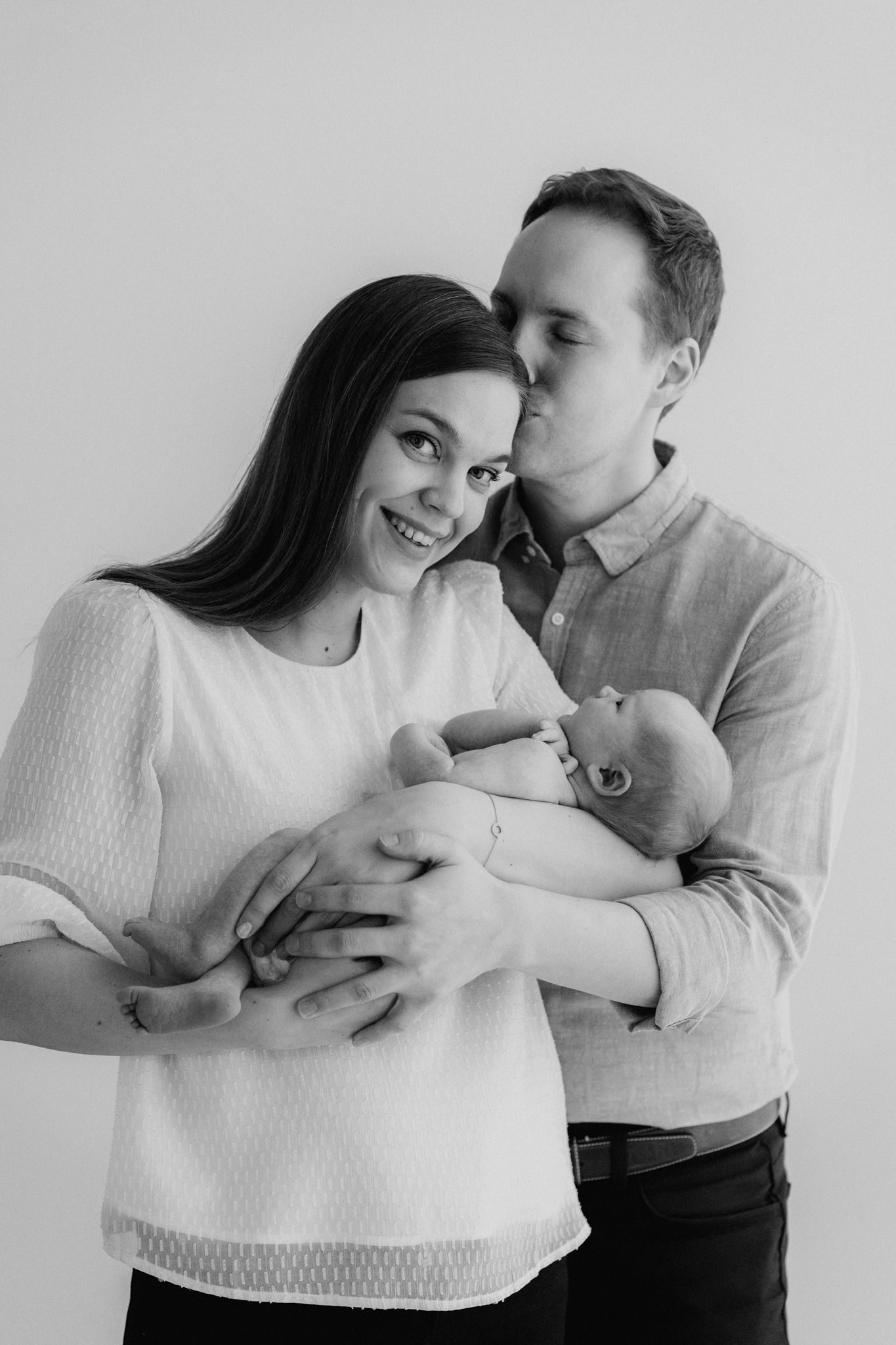 Black and white photo of smiling couple with man kissing womans cheek while holding newborn.