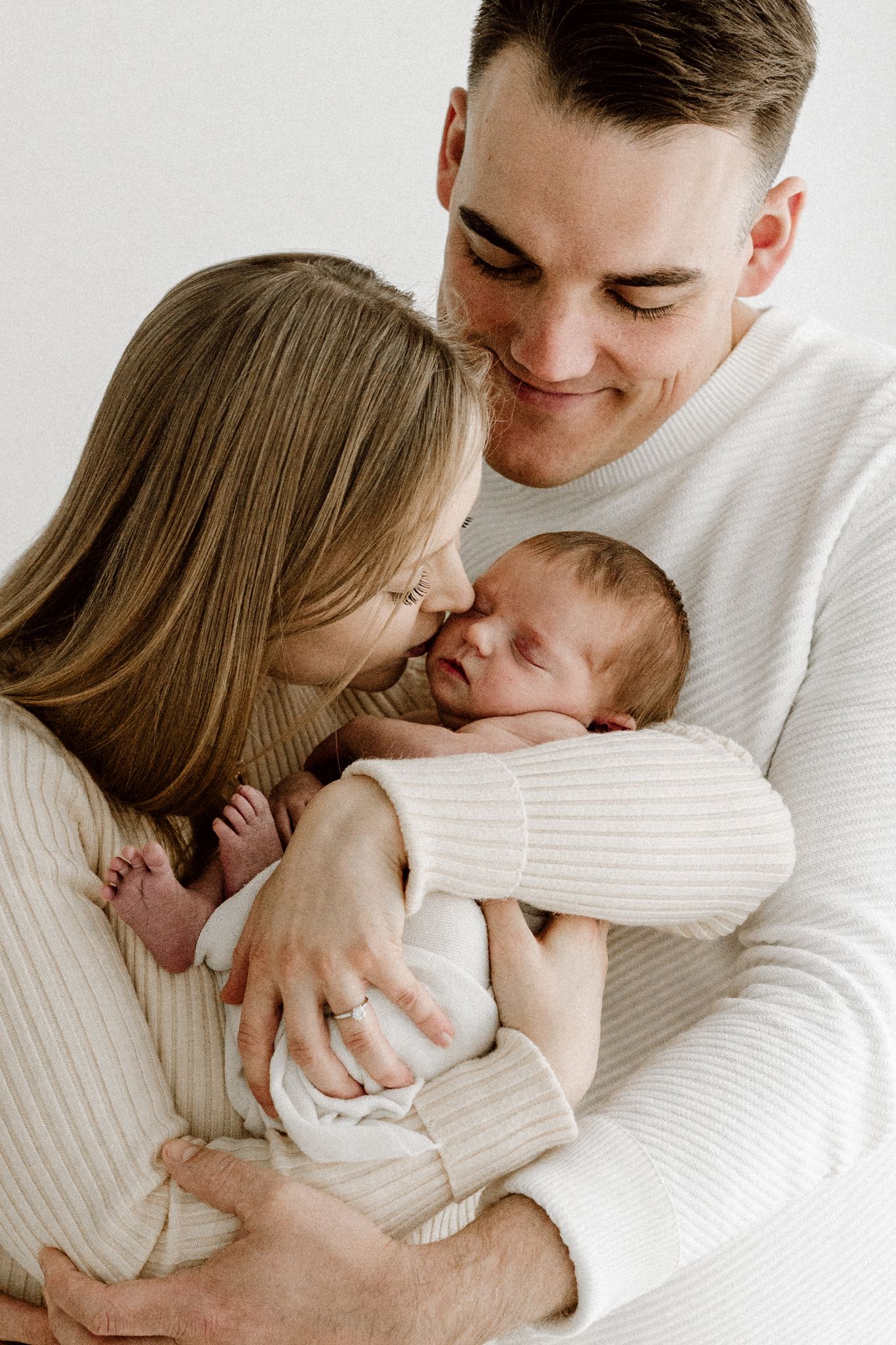 Young couple in white sweaters tenderly kissing their sleeping newborn baby together.