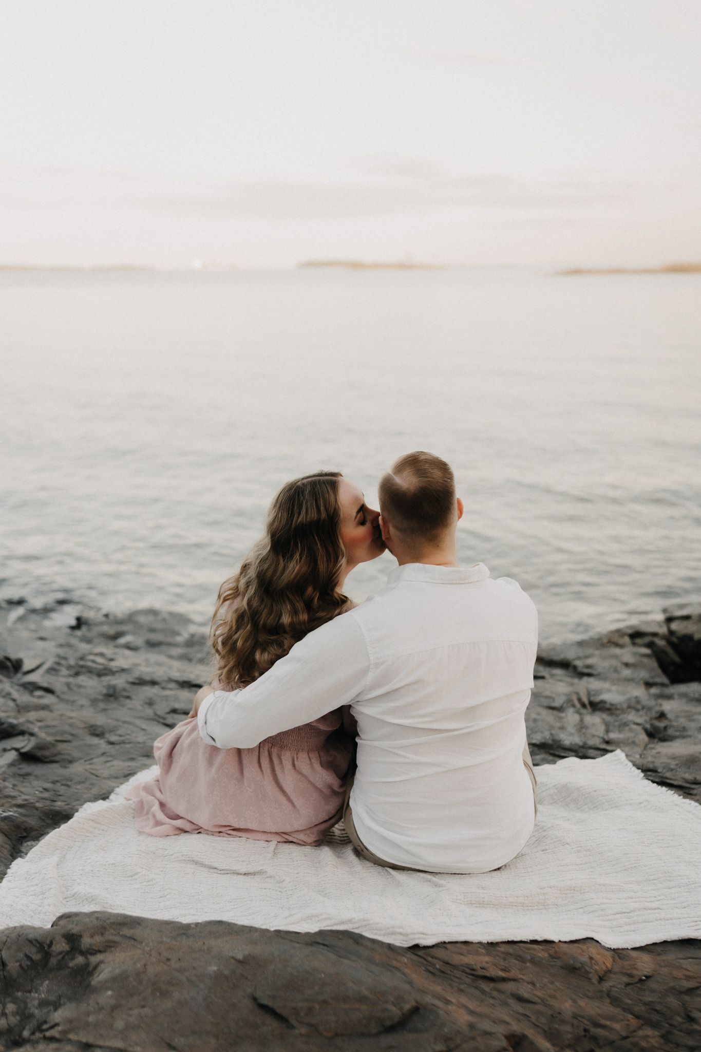 Couple sharing a kiss while sitting on white blanket facing calm lake waters at dusk.