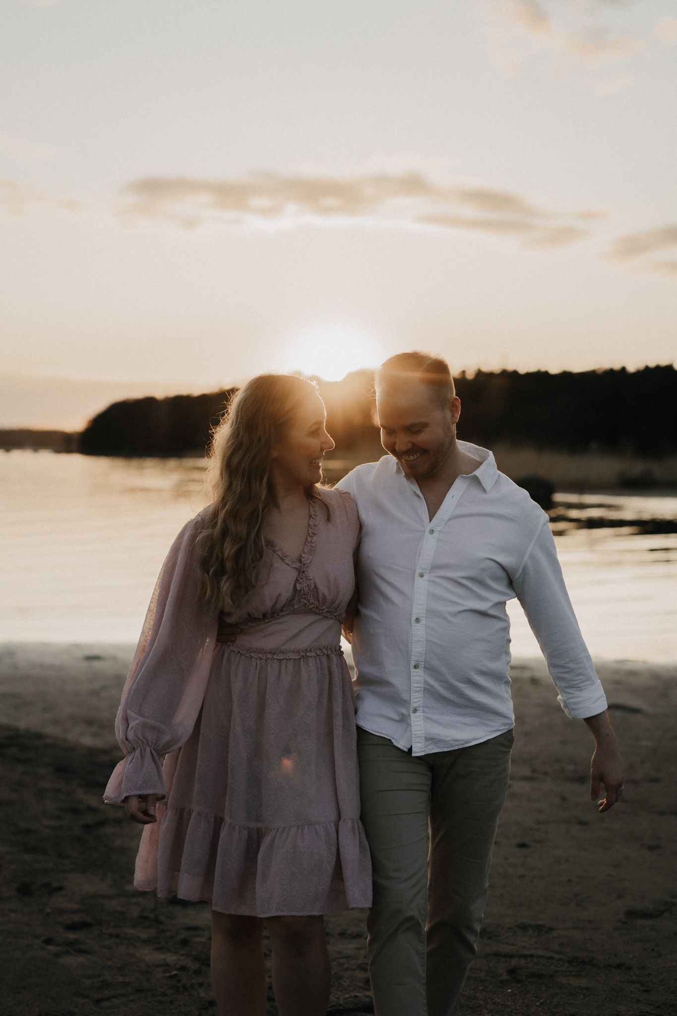 Couple walking on beach at sunset, woman in pink dress with man in white shirt embracing romantically.