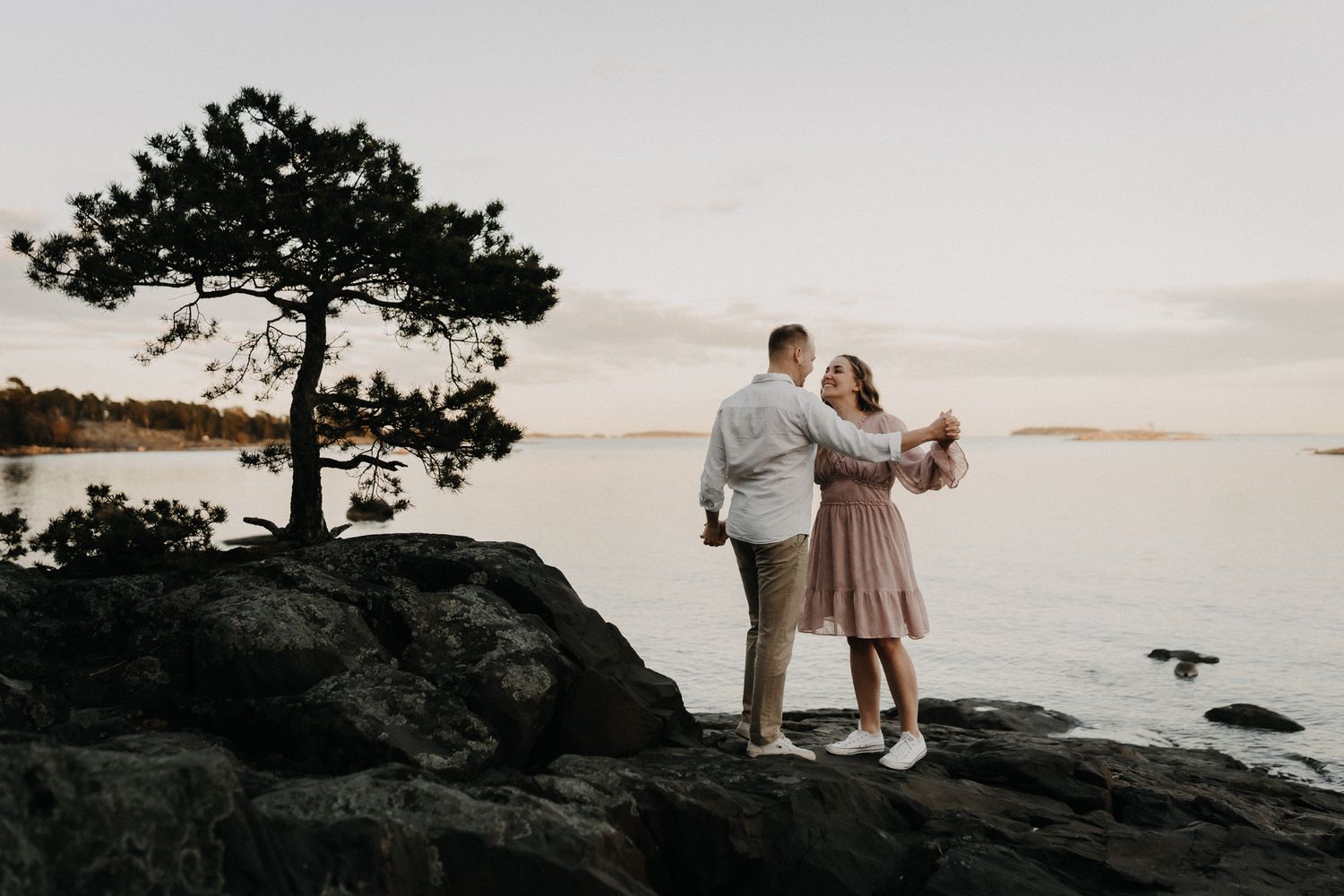 Romantic couple embraces near a lone pine tree on rocky coastal cliffs during a golden hour sunset.