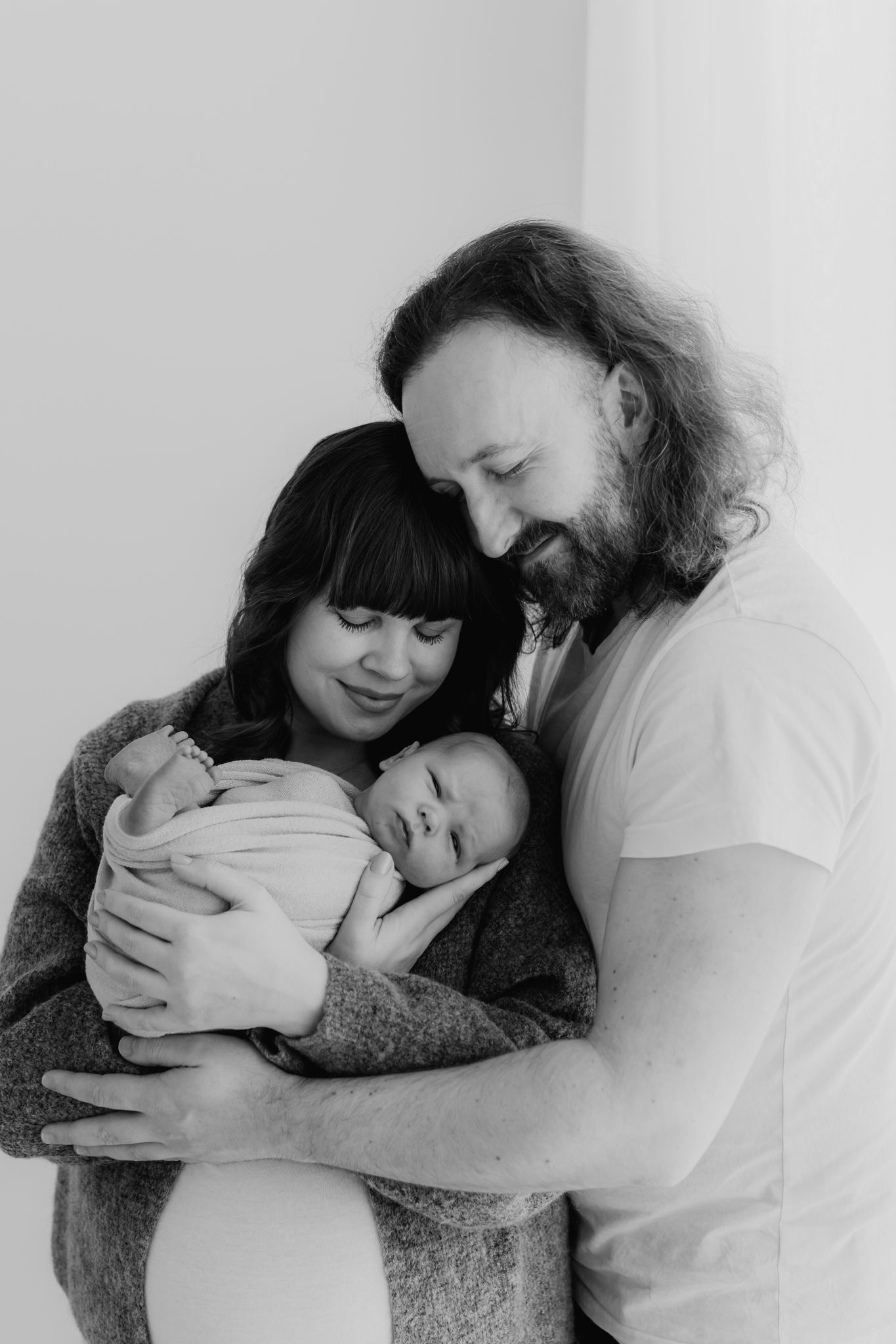 Black and white portrait of smiling parents with long hair lovingly holding their newborn baby.