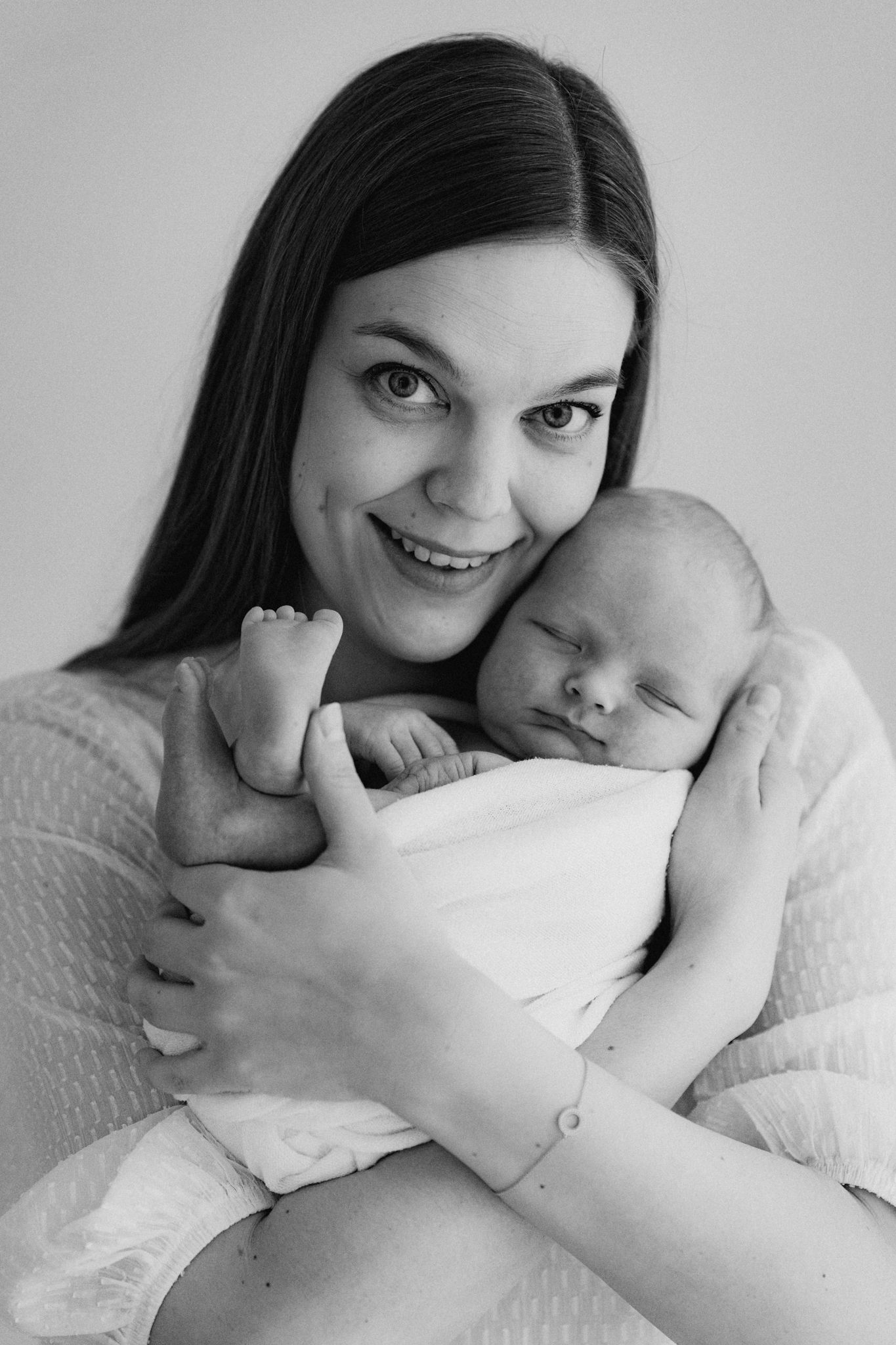 Black and white portrait of smiling mother holding sleeping swaddled newborn against her cheek.