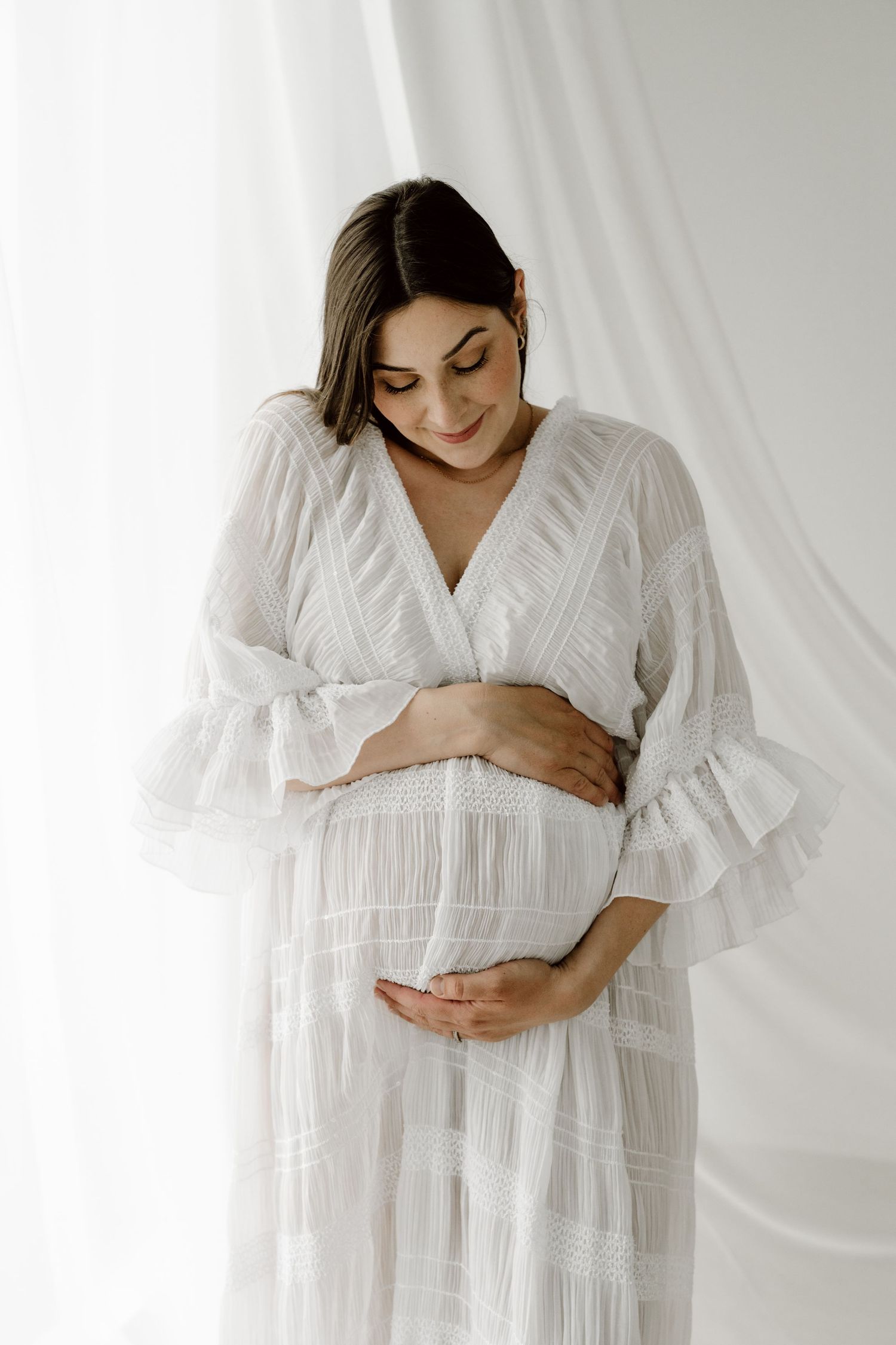 A smiling pregnant woman in a white lace dress lovingly holds her baby bump near white curtains.