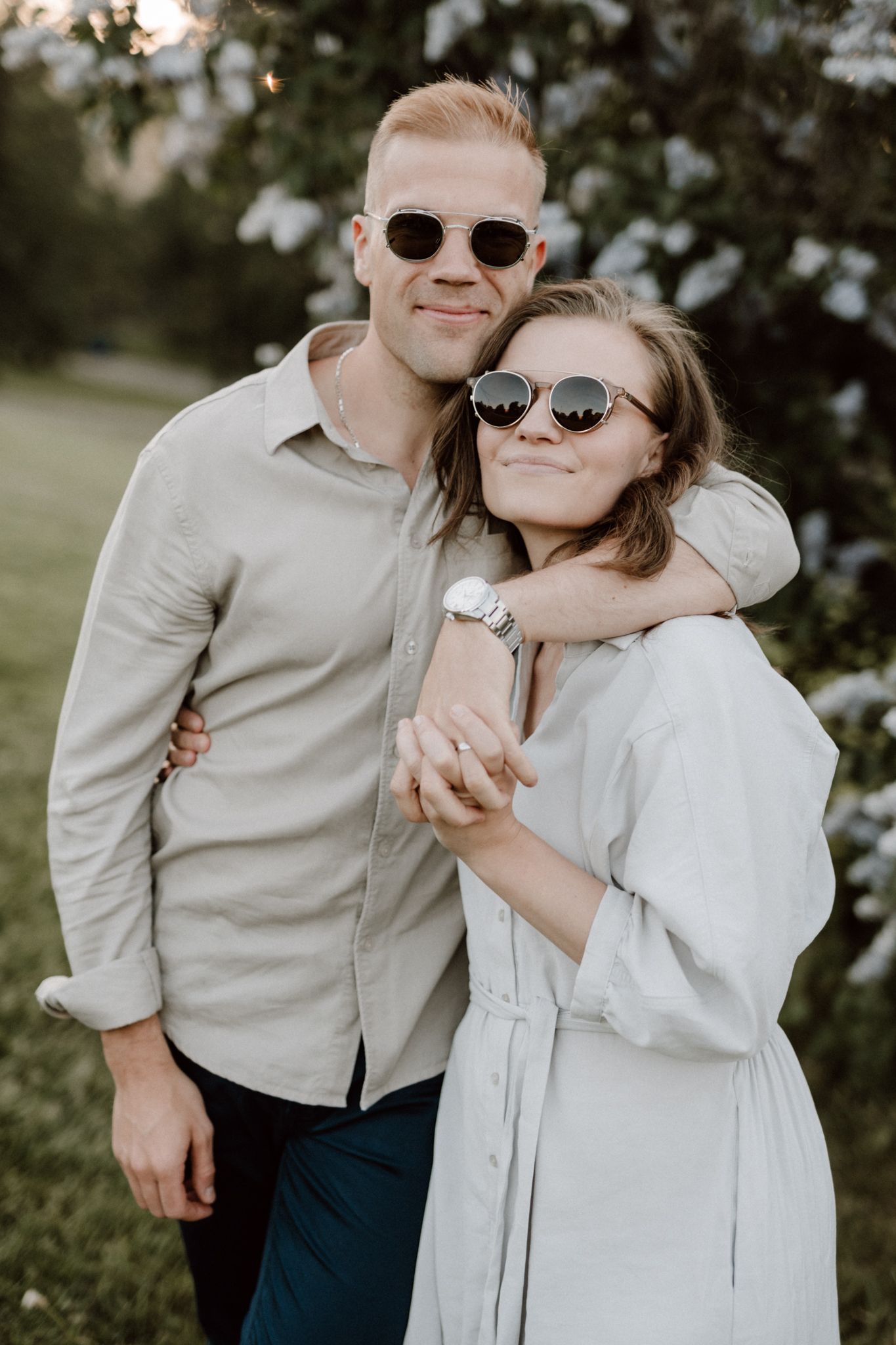 Smiling couple in sunglasses embracing near blooming white flowers at golden hour.