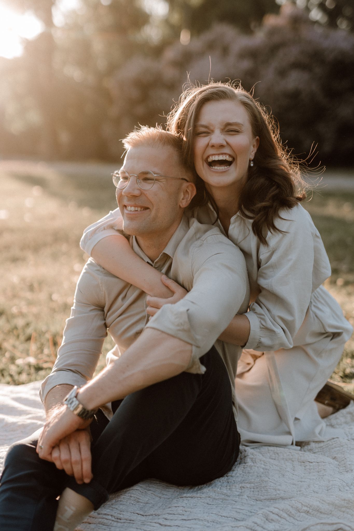 Laughing couple sitting on a blanket in a sunny park, woman hugging man from behind.