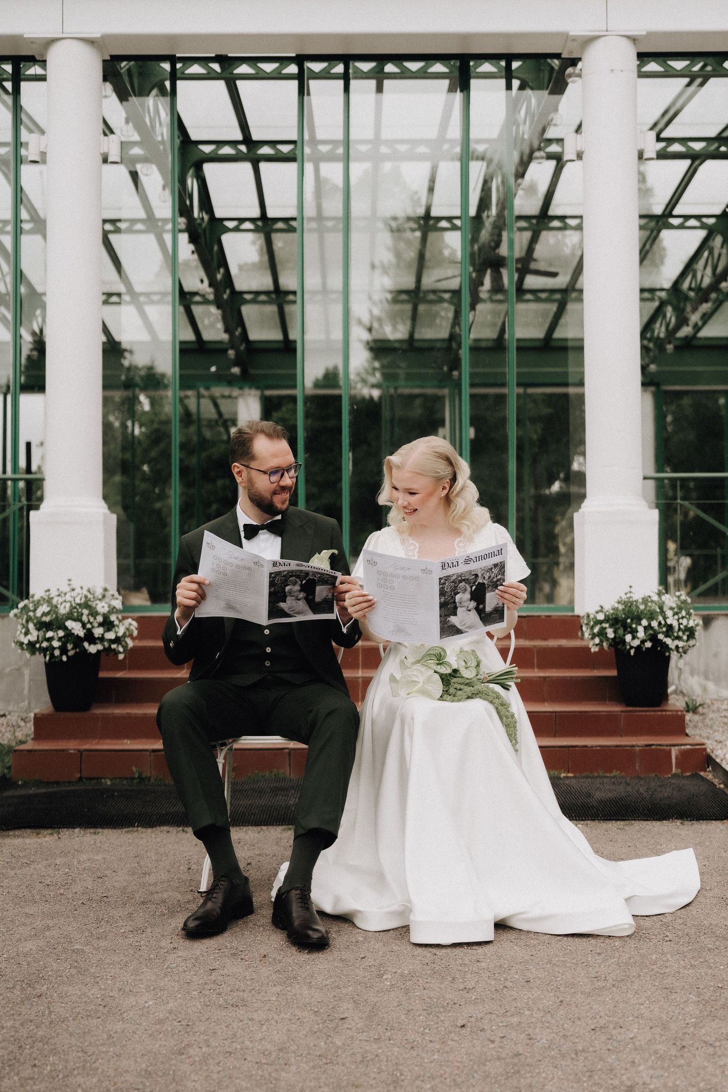 Bride and groom sit on steps reading wedding newspapers outside a greenhouse venue.