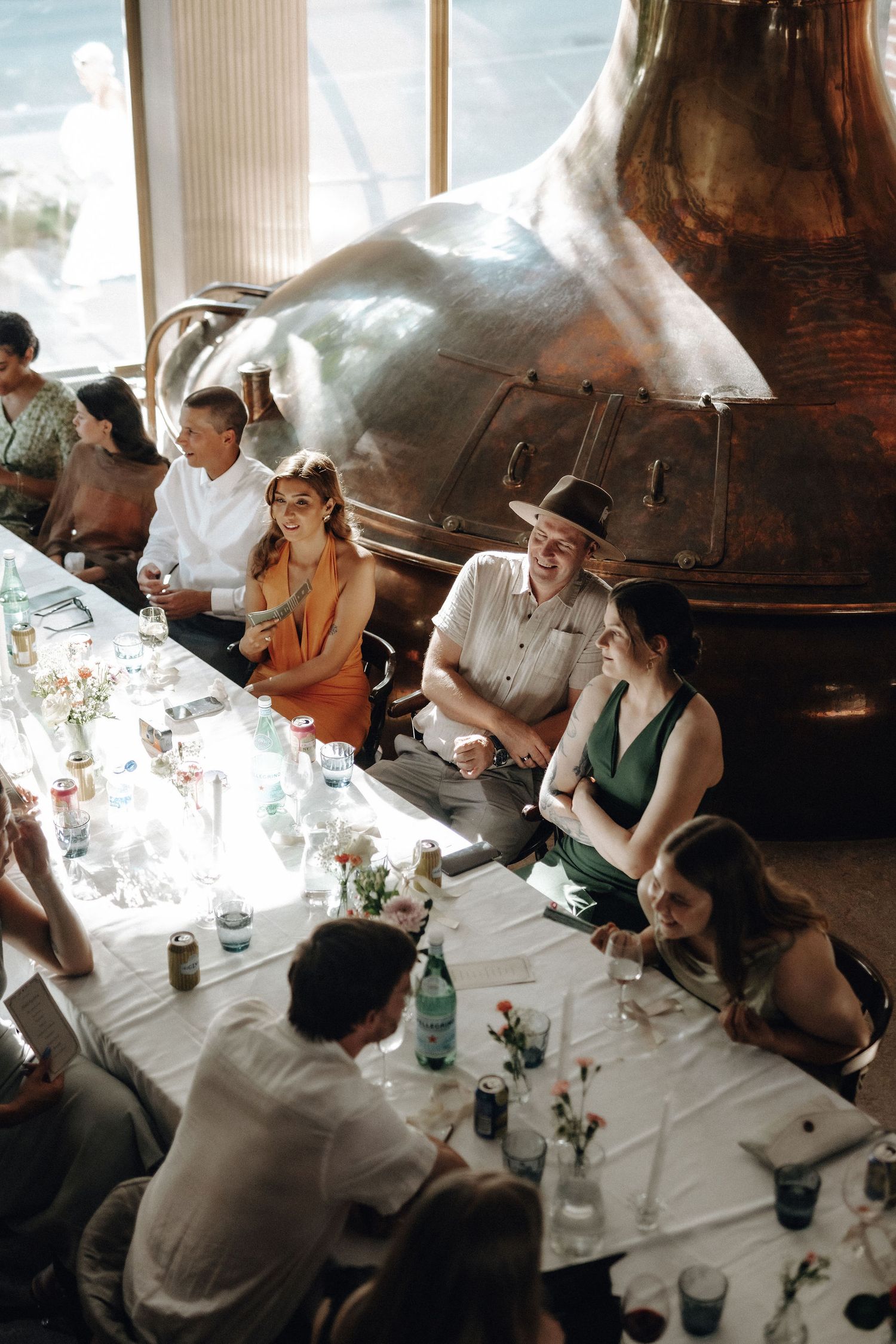 Wedding guests socializing at long reception table beside large copper brewery distillery tank.