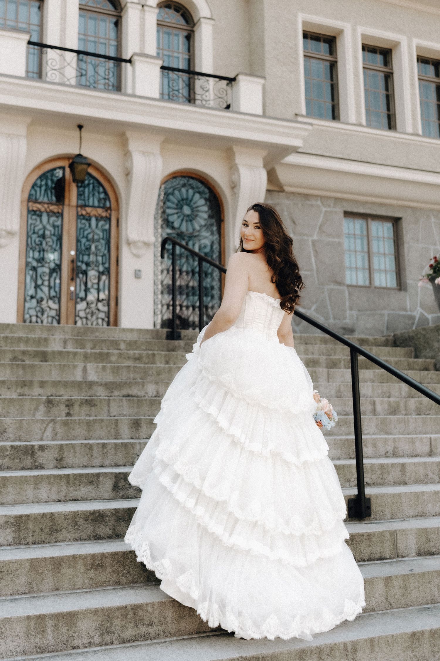 Bride in a voluminous tiered white strapless gown ascending stone steps of a classical building.