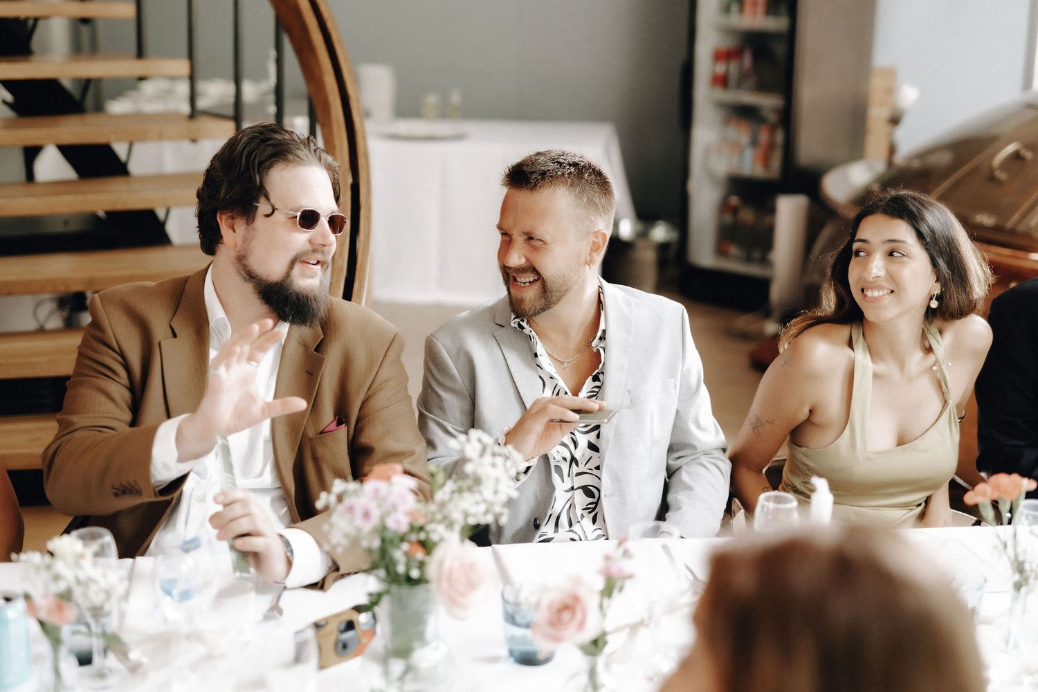 Three wedding guests laughing and conversing at a floral-decorated reception dinner table.