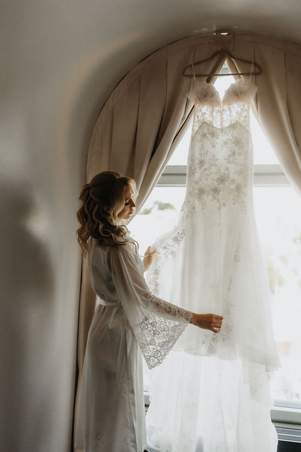 Bride in white lace robe admires her wedding gown hanging in arched window before ceremony.