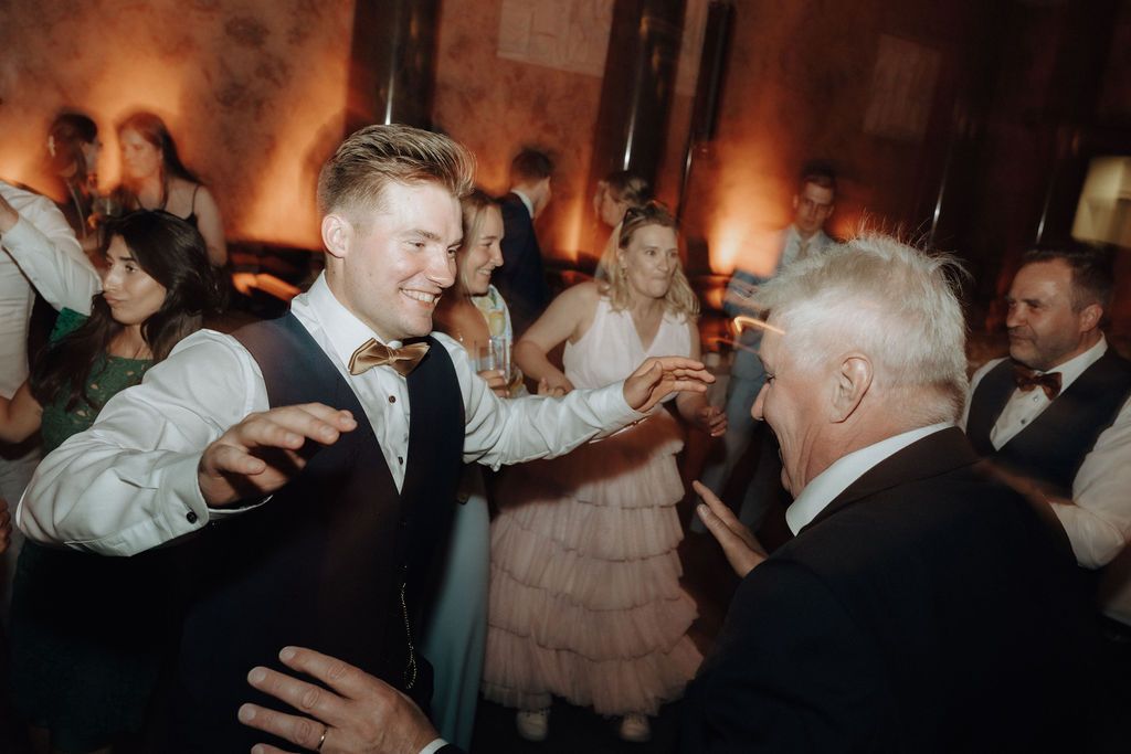 Smiling groom in vest and bow tie dancing with an older guest at a wedding reception.