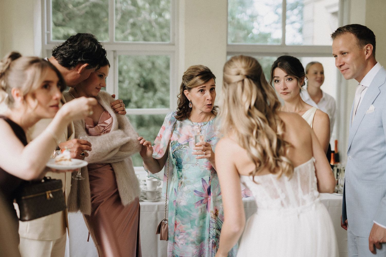 Bride in white gown conversing with elegantly dressed wedding guests in a bright reception hall.