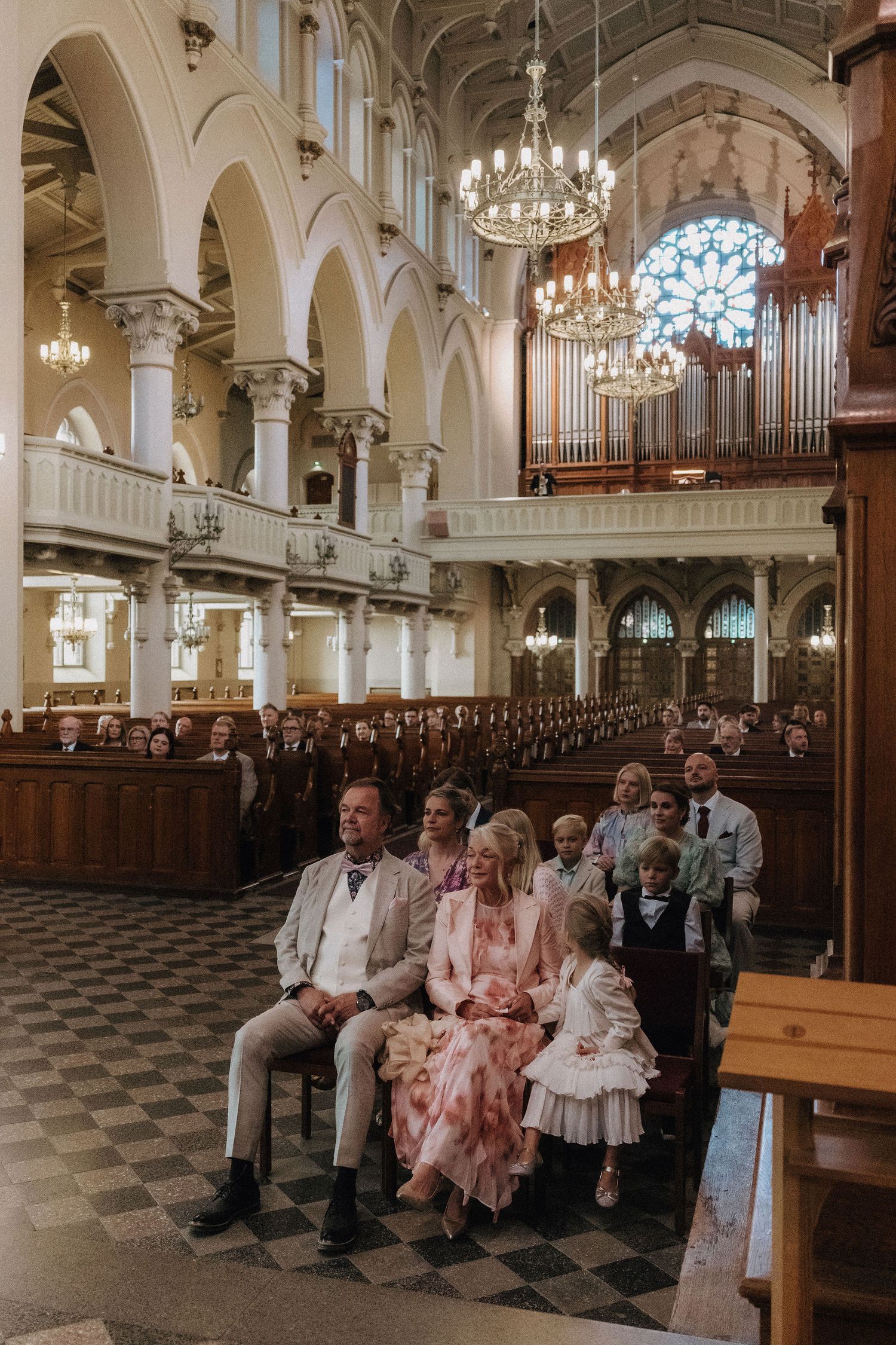 Wedding guests fill ornate church pews beneath grand chandeliers and large pipe organ.