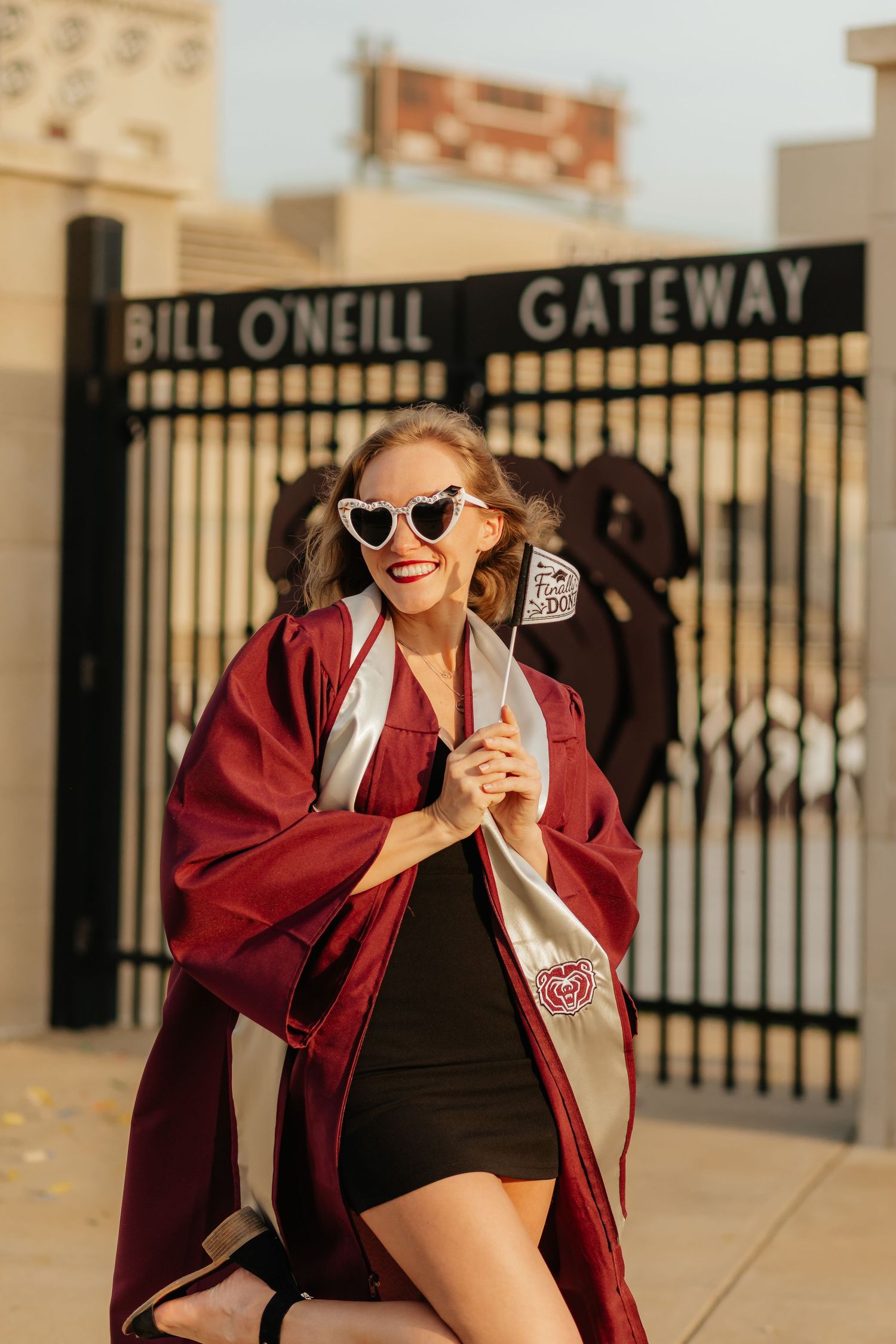 MSU grad in gown in front of Bill ONeill gate holding a congrats grad flag