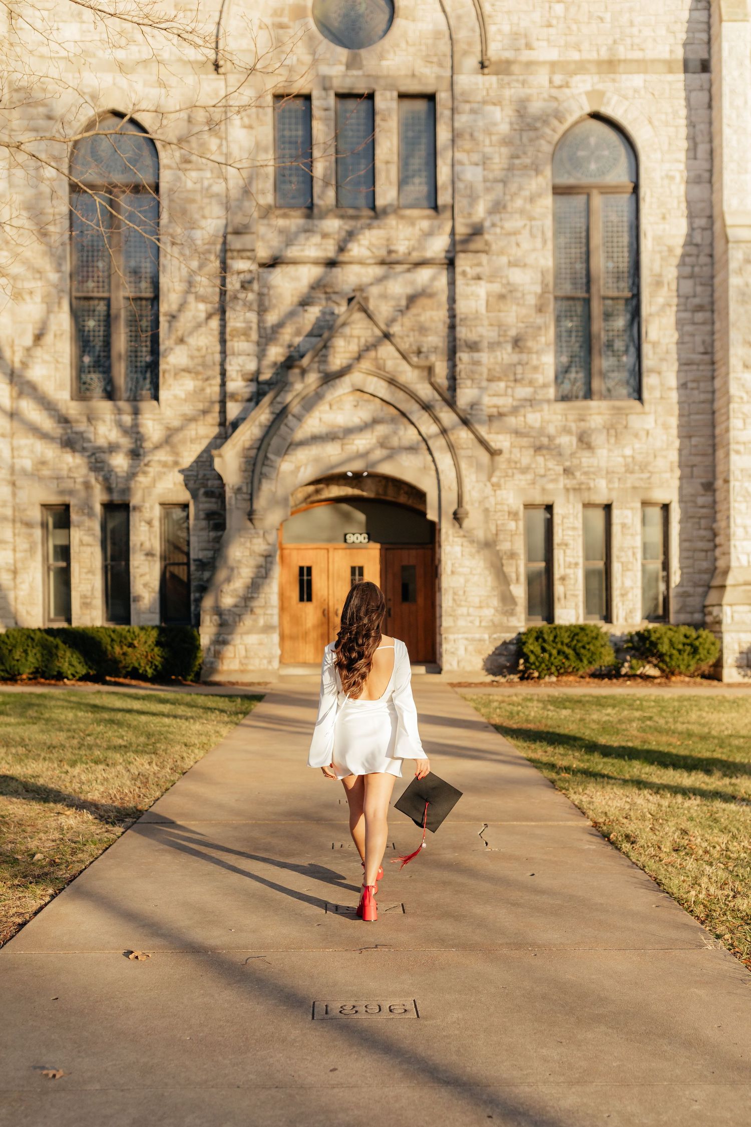 Drury grad walking away from camera holding cap wearing white dress and red heels