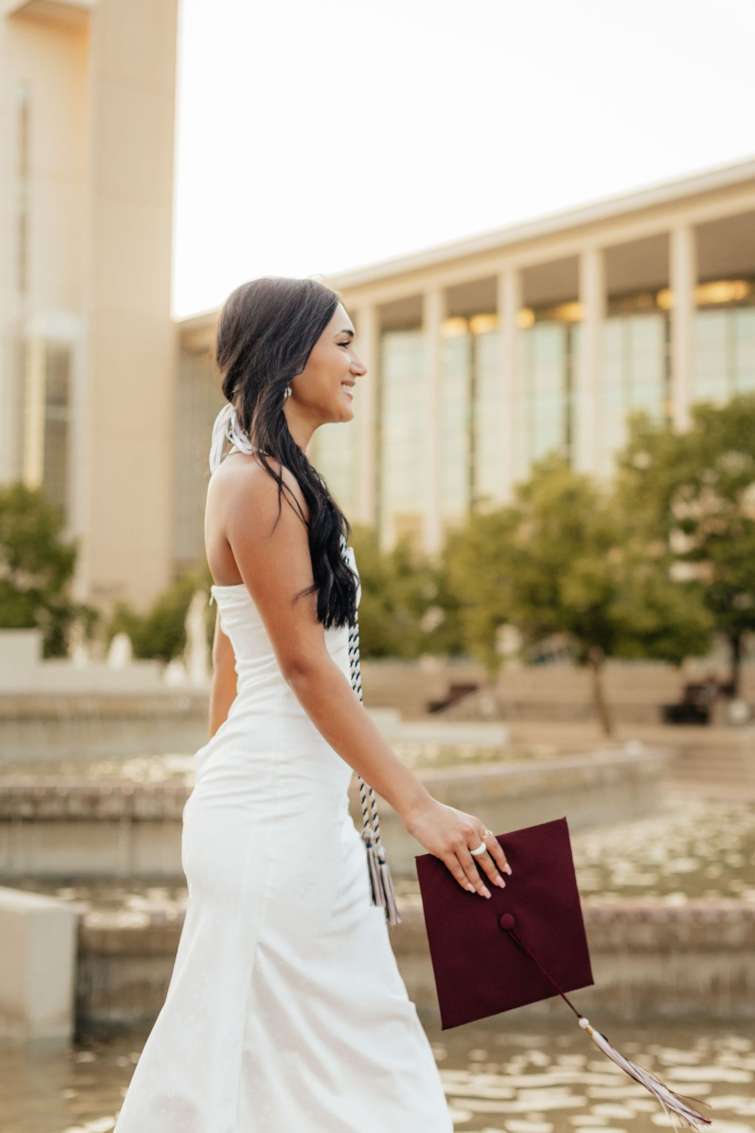 woman in white silk dress holding grad cap walking