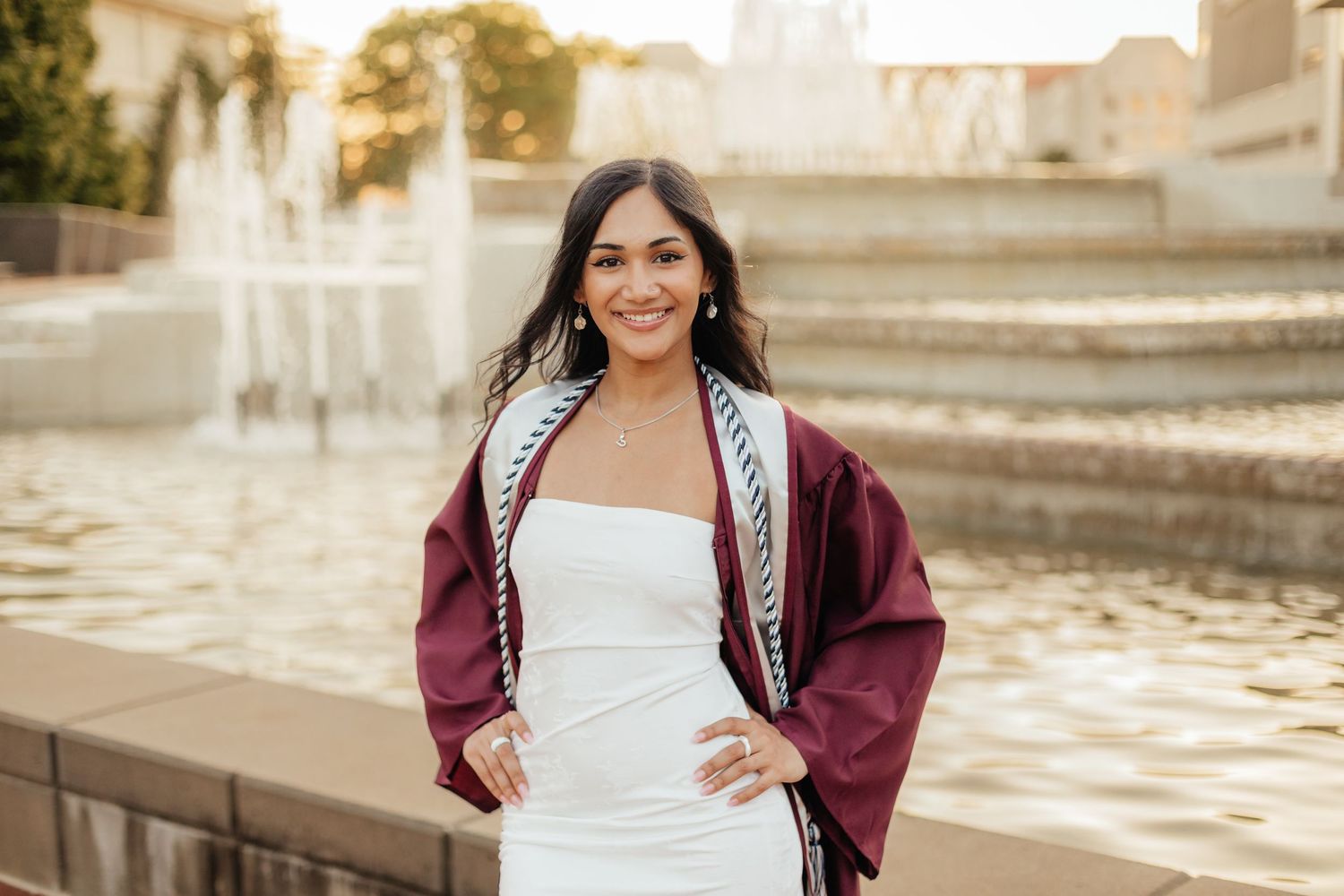 MSU grad wearing gown, academic cords, and stole standing in front of fountain