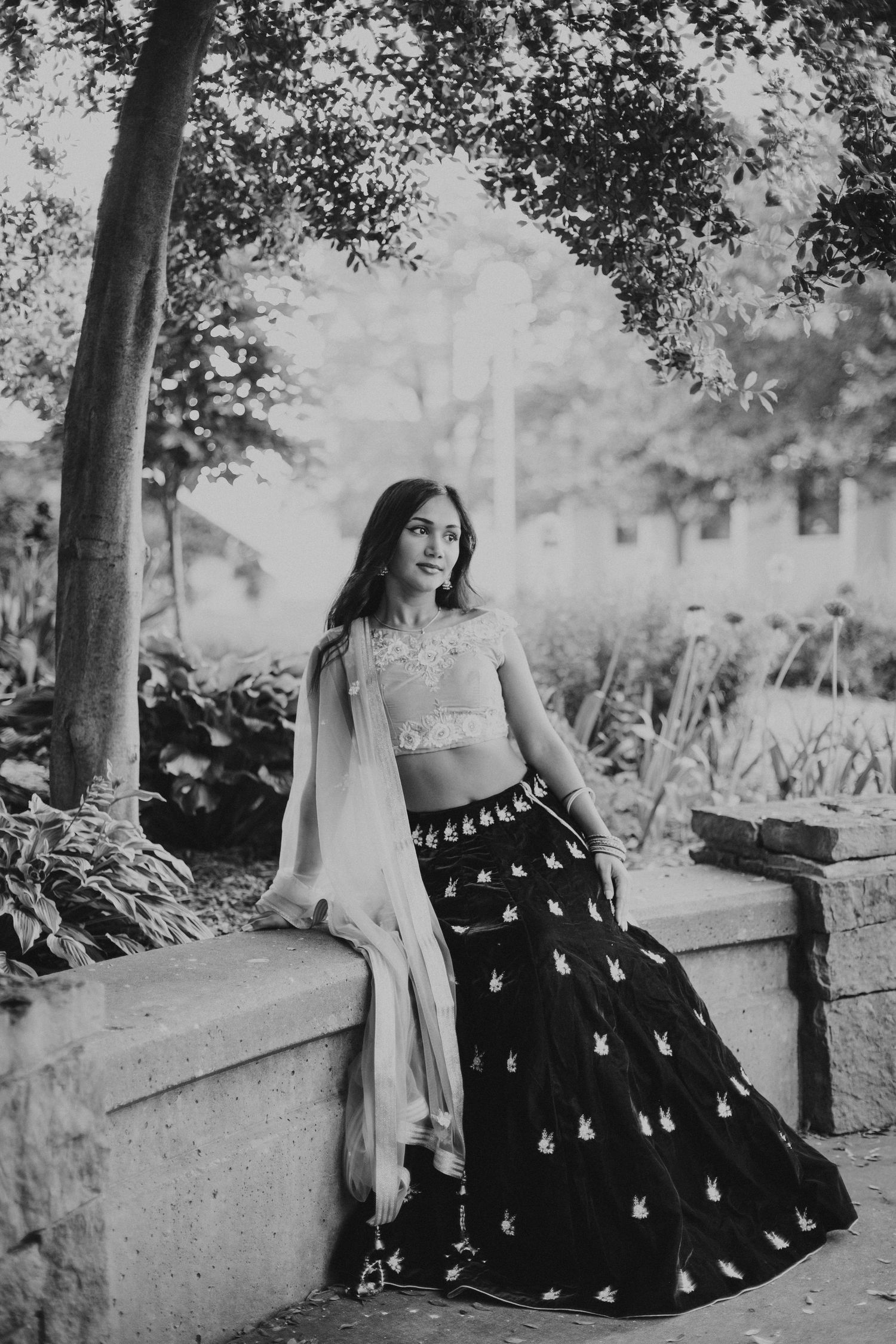 black and white photo of woman sitting on a ledge looking away wearing a lehenga