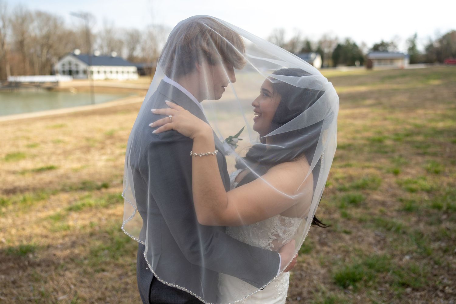 A couple shares an intimate moment under a sheer wedding veil in an outdoor field setting.