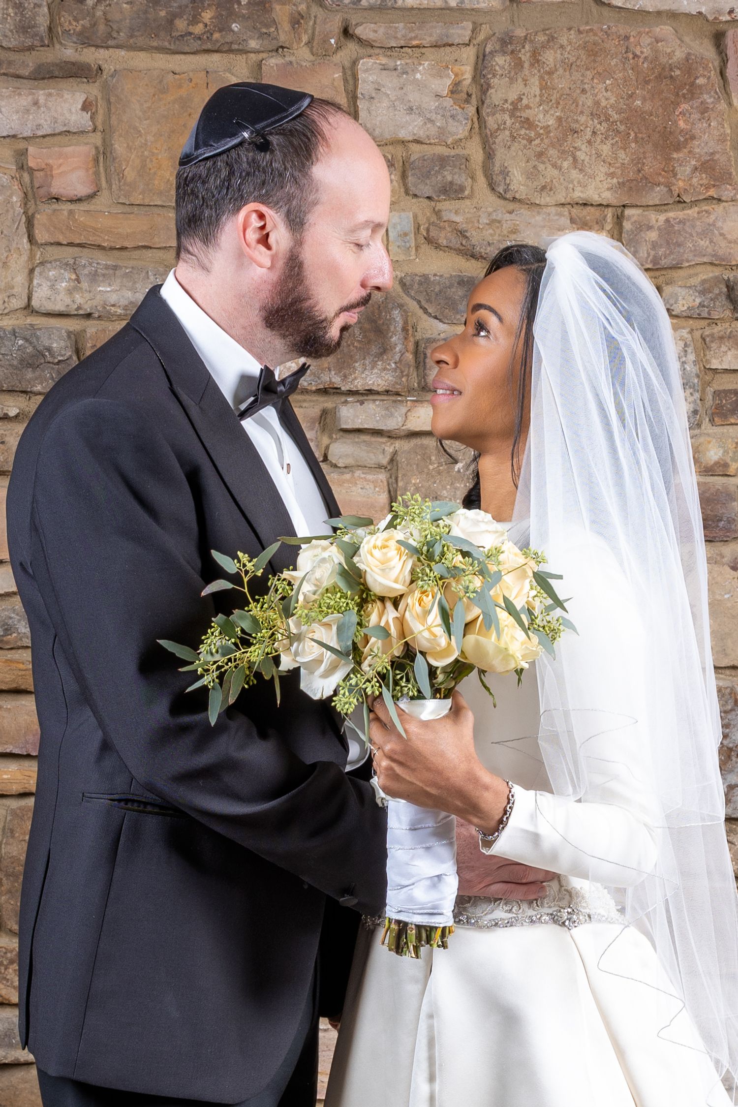Bride and groom share intimate moment against stone wall with elegant floral bouquet.