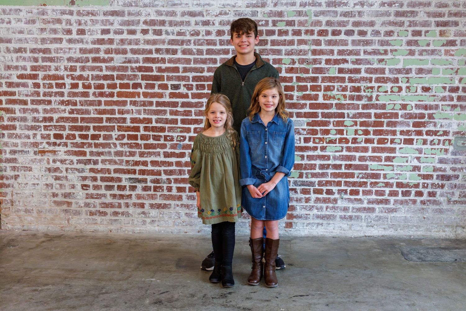 Three children stand against a weathered brick wall in matching winter coats and boots.