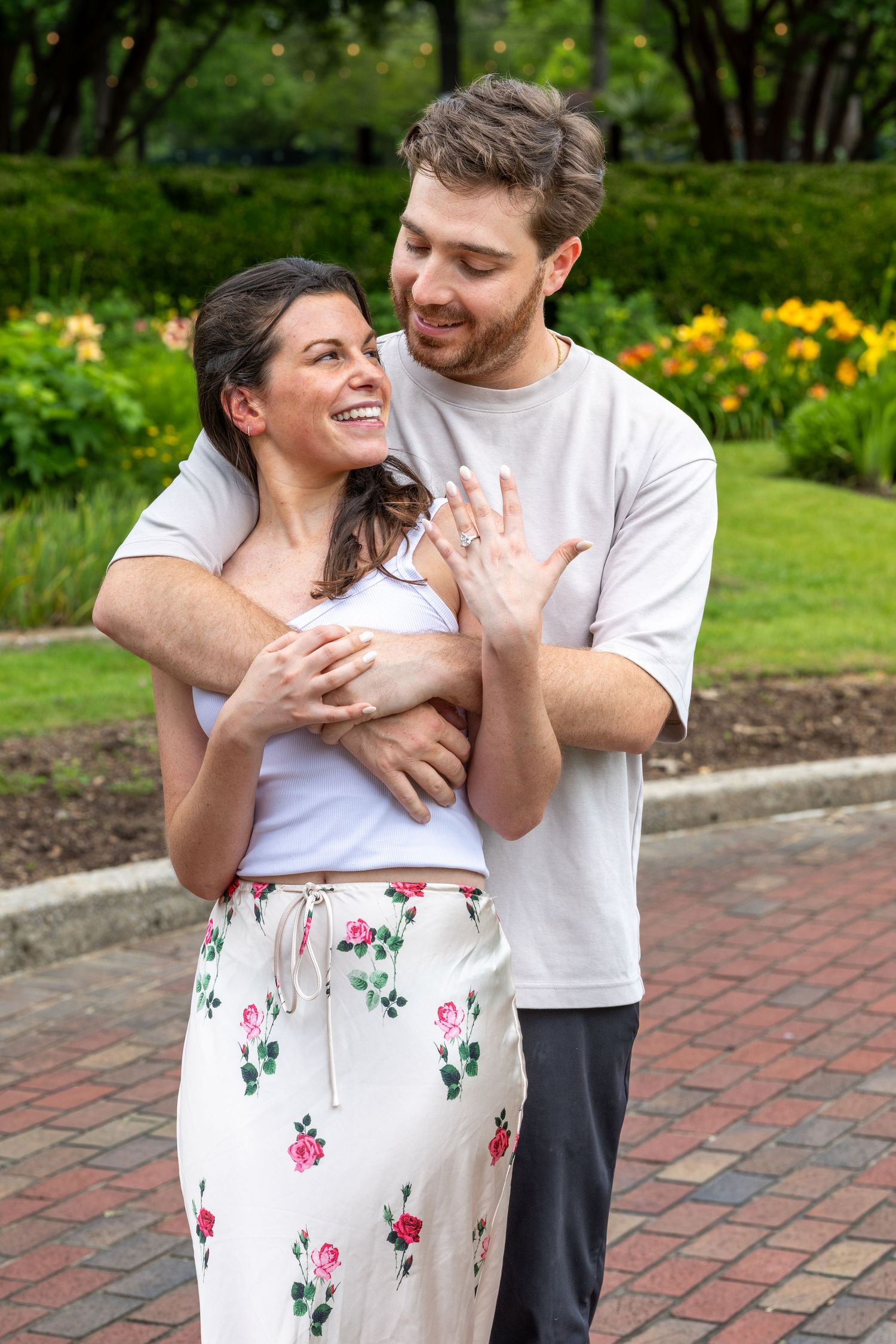 Happy couple embracing in a garden park, woman showing off engagement ring while smiling joyfully.