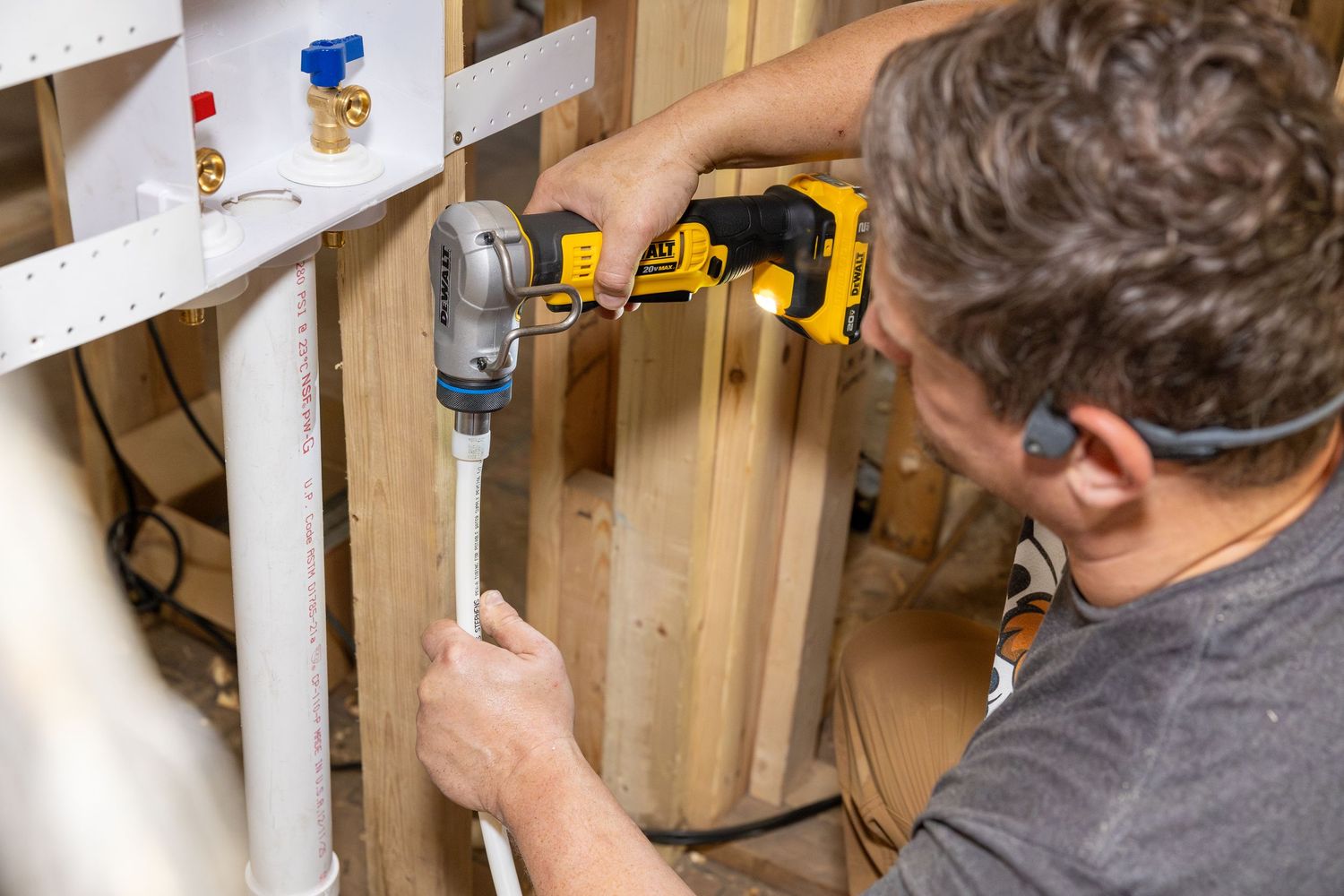 Plumber using a DeWalt drill to install white PVC pipe on a water heater in a framed wall.