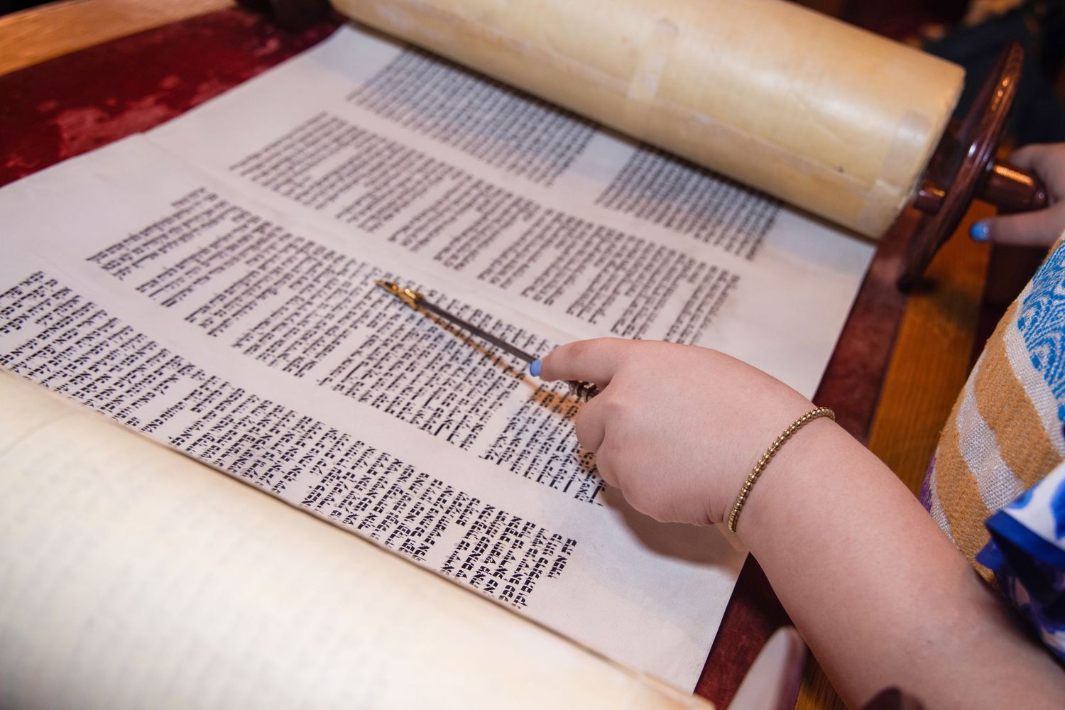 A hand uses a pointer tool to read text from an open Torah scroll during a religious service.