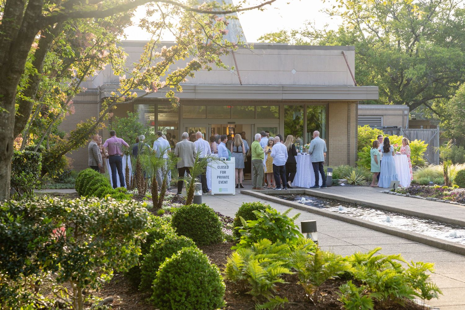 Guests gather outside a garden venue for an outdoor event surrounded by lush greenery and blooming trees in warm sunlight.