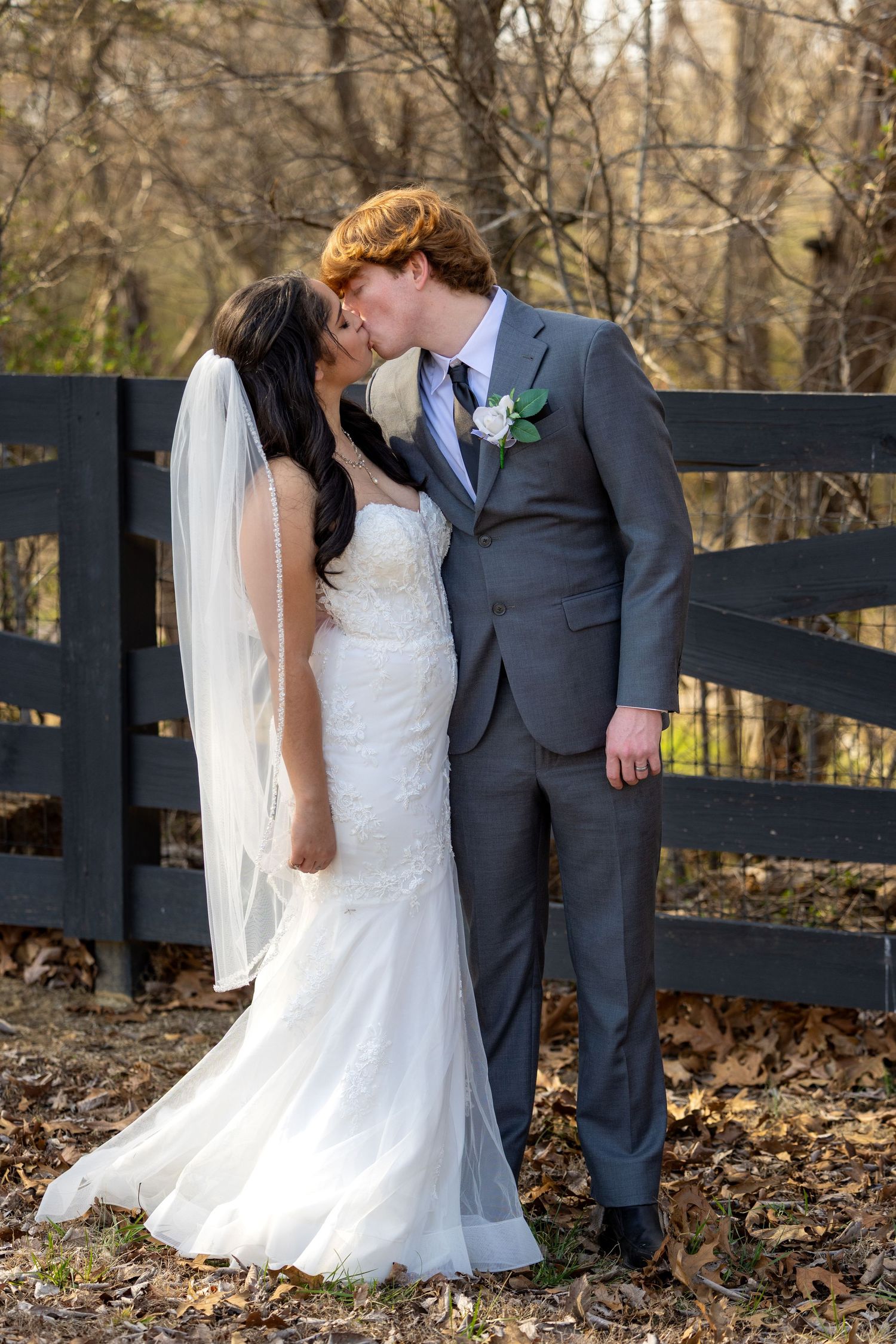 A bride and groom share an intimate moment against a rustic black fence surrounded by fallen autumn leaves.