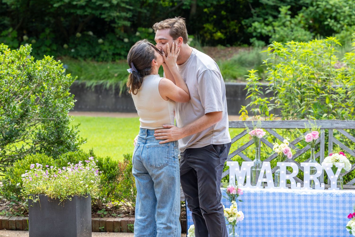 Couple sharing a romantic kiss near a MARRY sign during an outdoor marriage proposal in a lush green park.