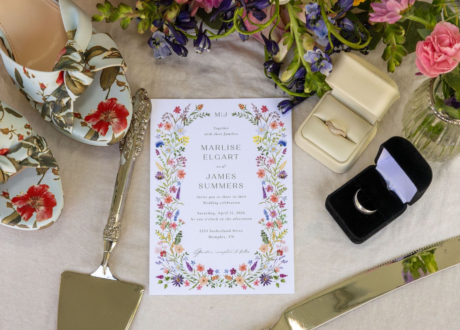 Elegant wedding invitation with floral border surrounded by rings, floral heels, gold cake servers, and fresh flowers.