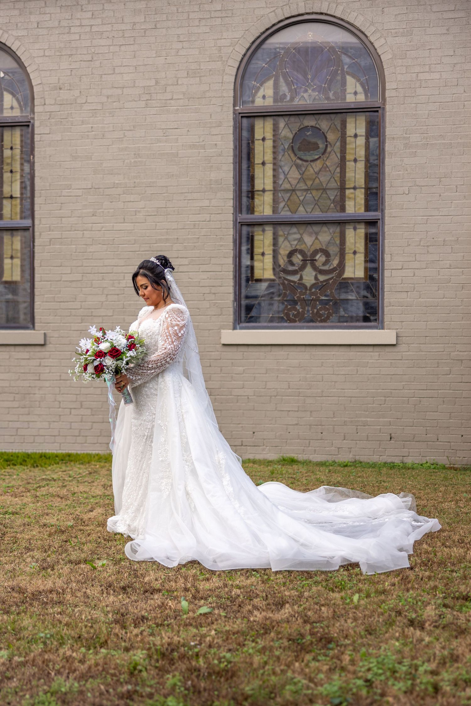Bride in flowing white lace gown poses against church exterior with stained glass windows.
