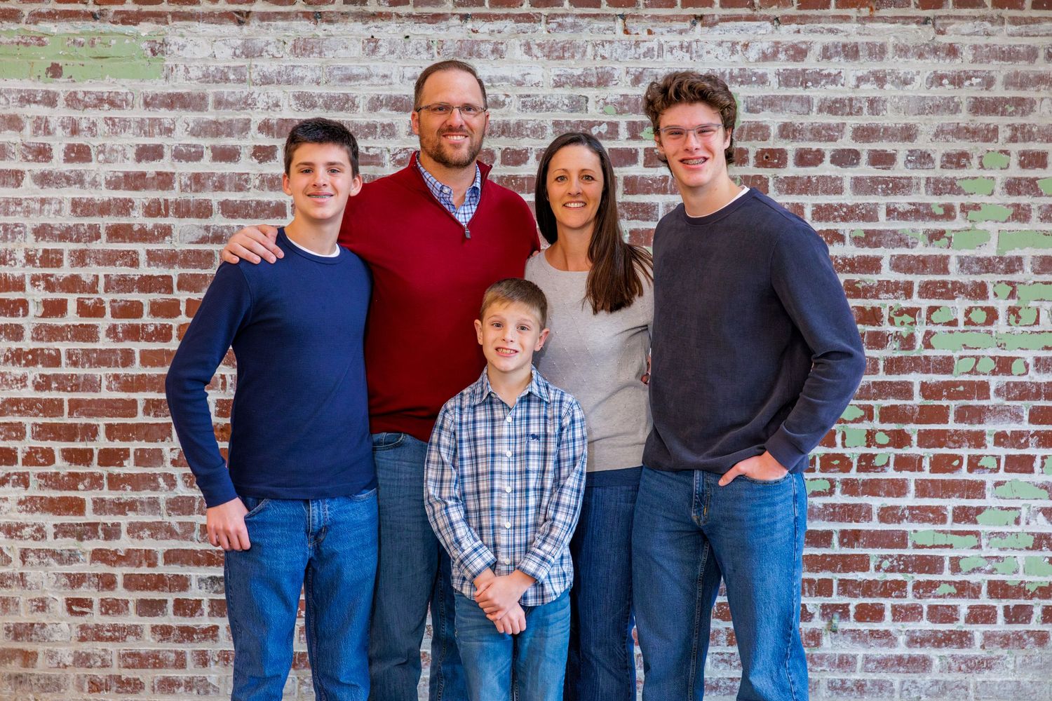 A family group poses together in coordinated casual outfits against a weathered brick wall.