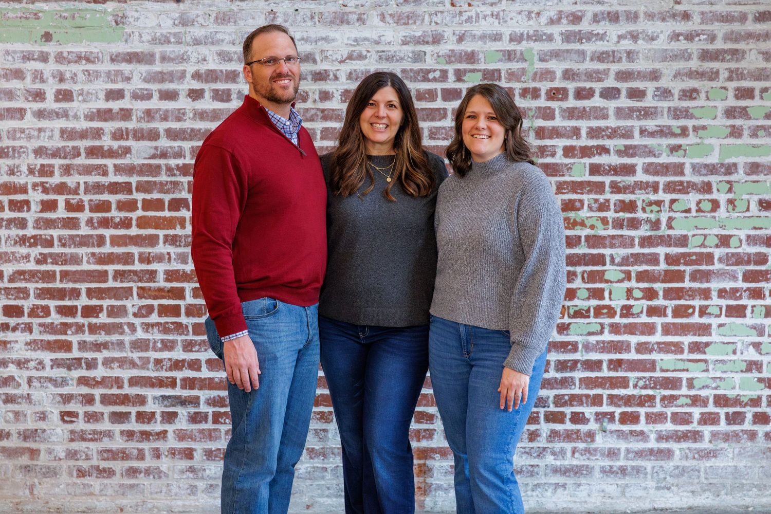 Family members pose together in front of a rustic brick wall wearing casual sweaters and jeans.