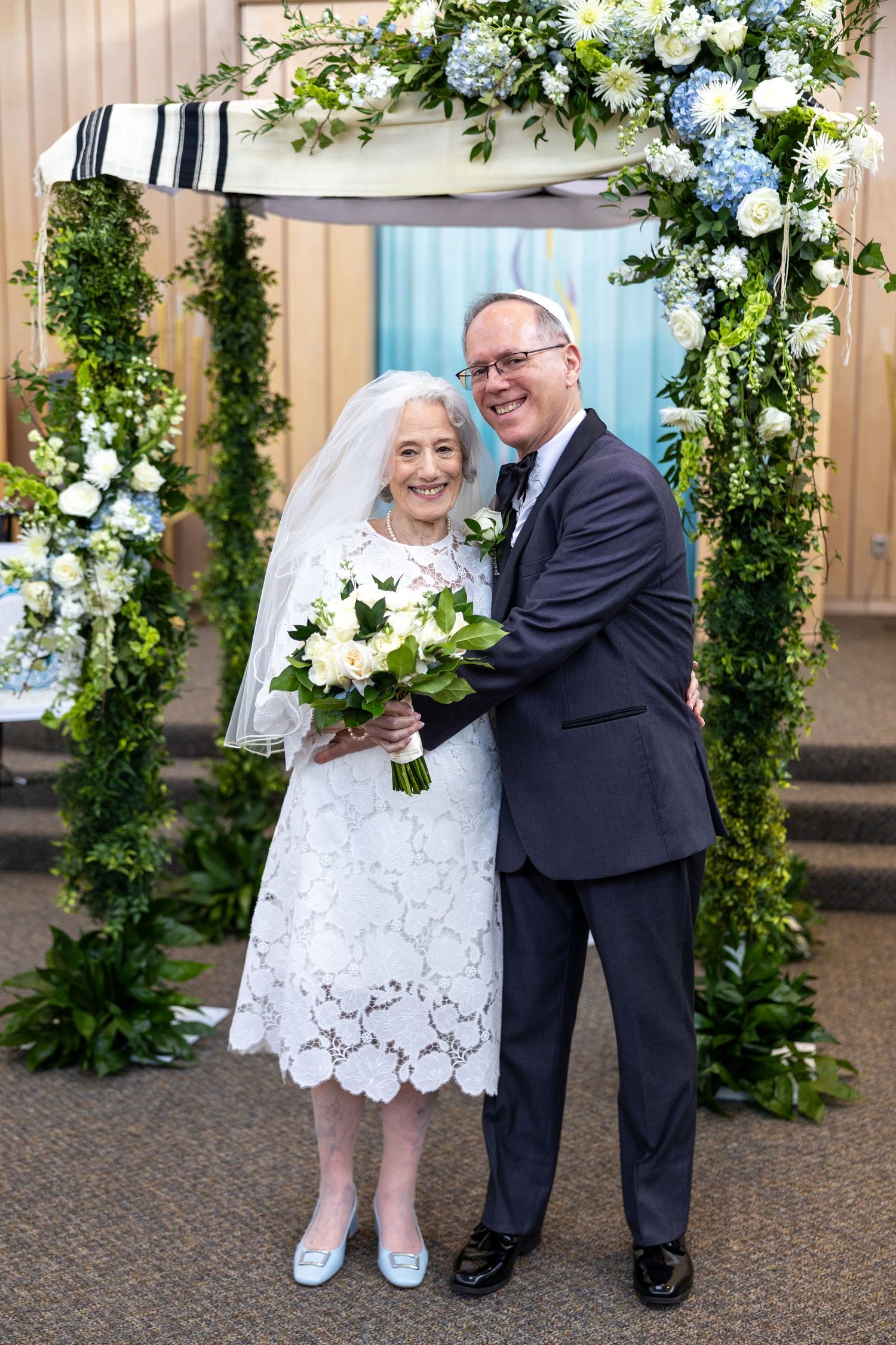 A couple poses under a floral wedding arch decorated with blue and white flowers and greenery.