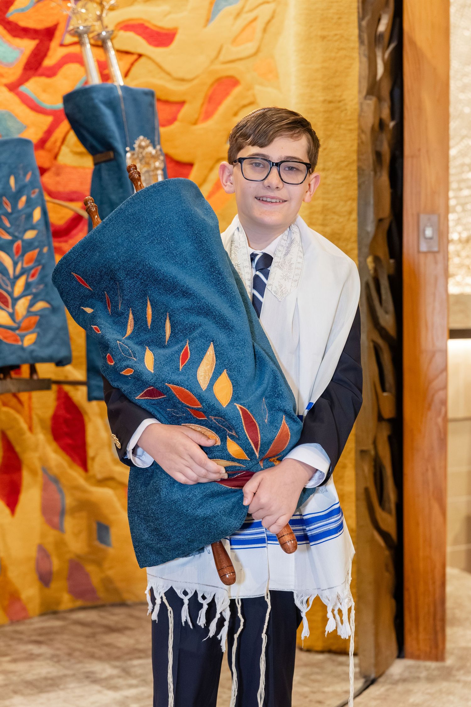 Young boy in tallit holds Torah scroll during Bar Mitzvah ceremony in decorated synagogue.
