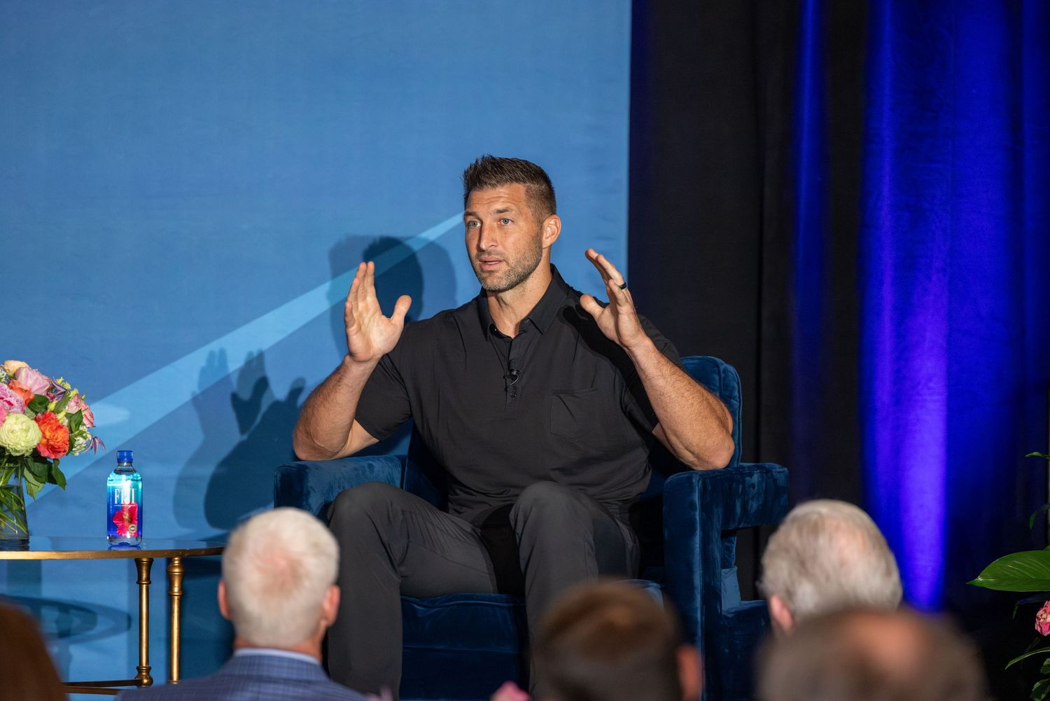 Man in black shirt gestures while speaking on stage at a conference, seated in a blue velvet chair.