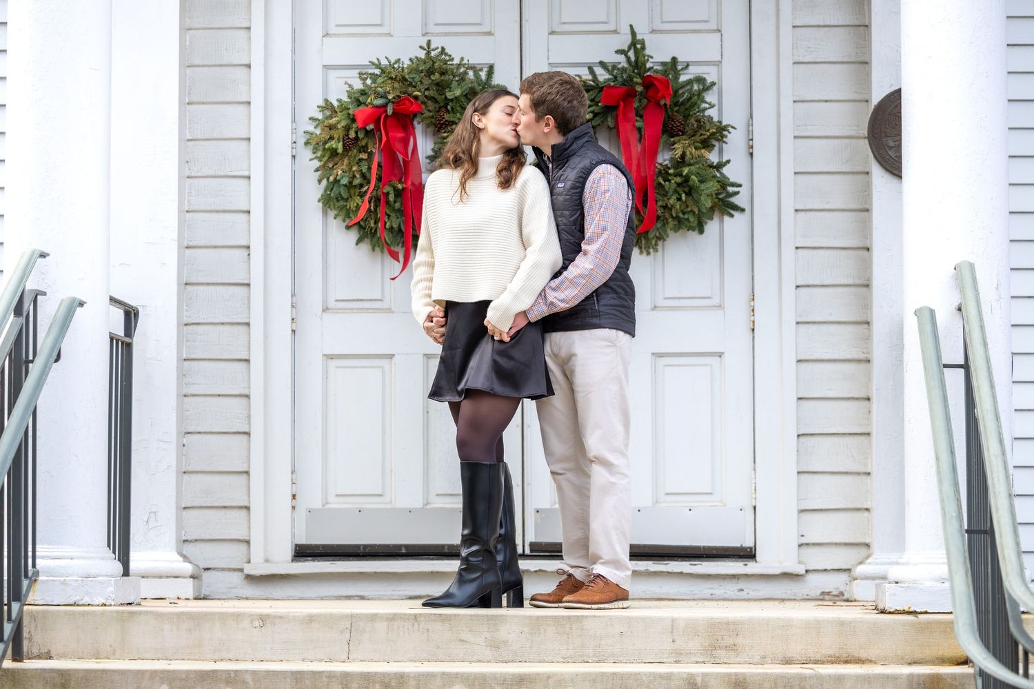 A couple stands close together on steps decorated with Christmas wreaths and red bows.