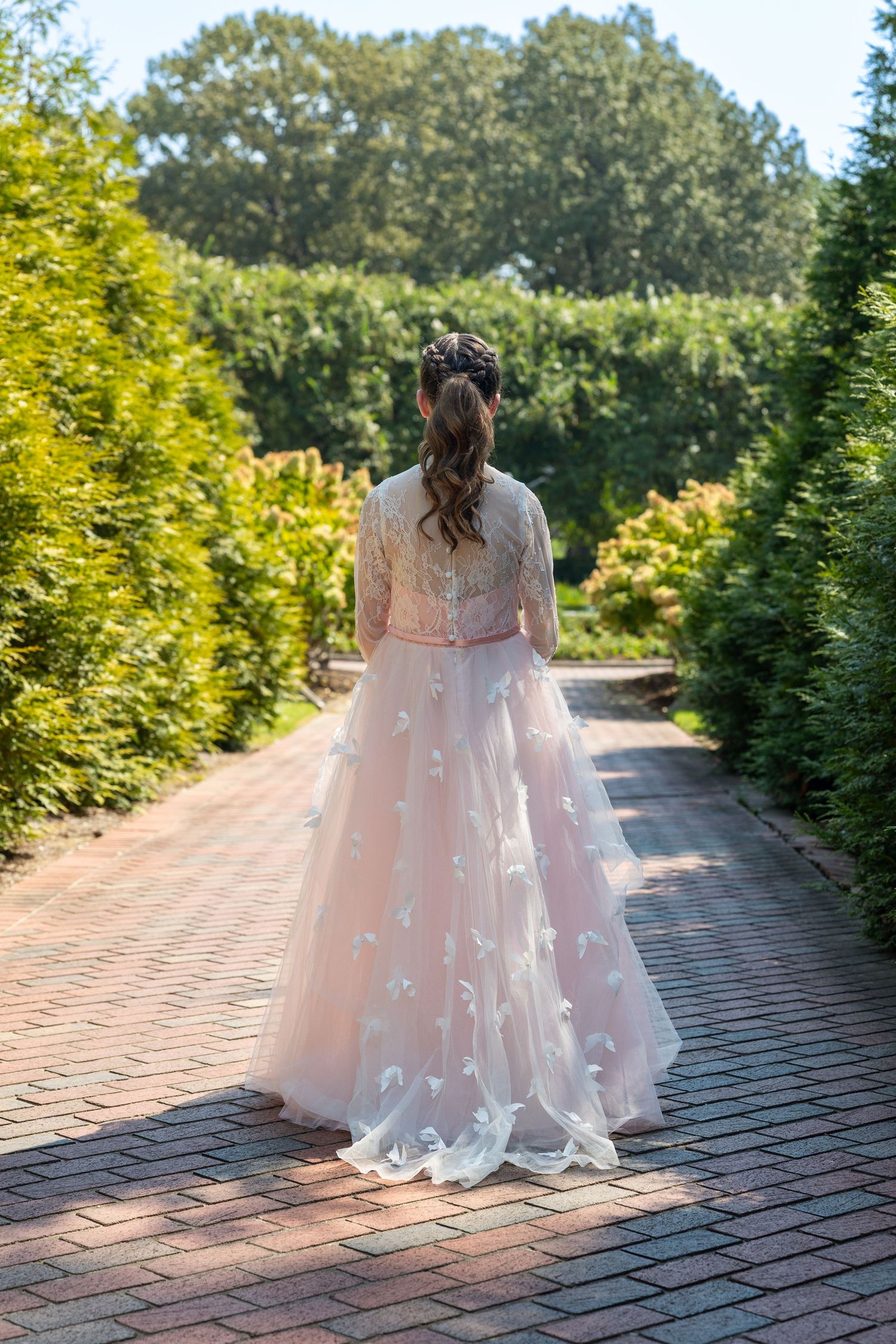Bride in pink floral ball gown with butterfly appliques walks garden brick path, back view, lush greenery surroundings.