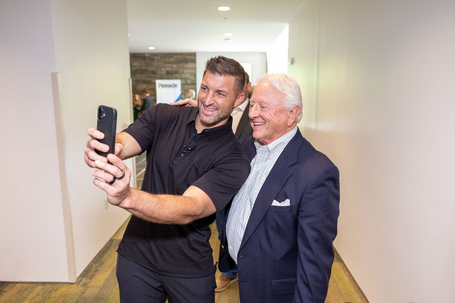 Two men take a selfie together in a modern hallway during a professional networking event.