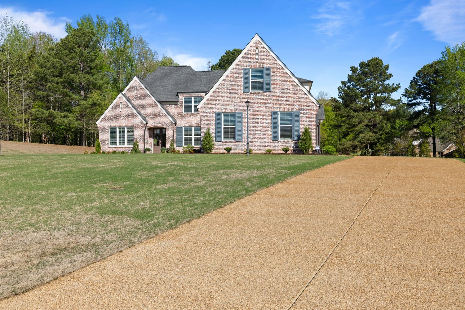 Large brick home with green lawn and wide concrete driveway surrounded by trees on a sunny day.